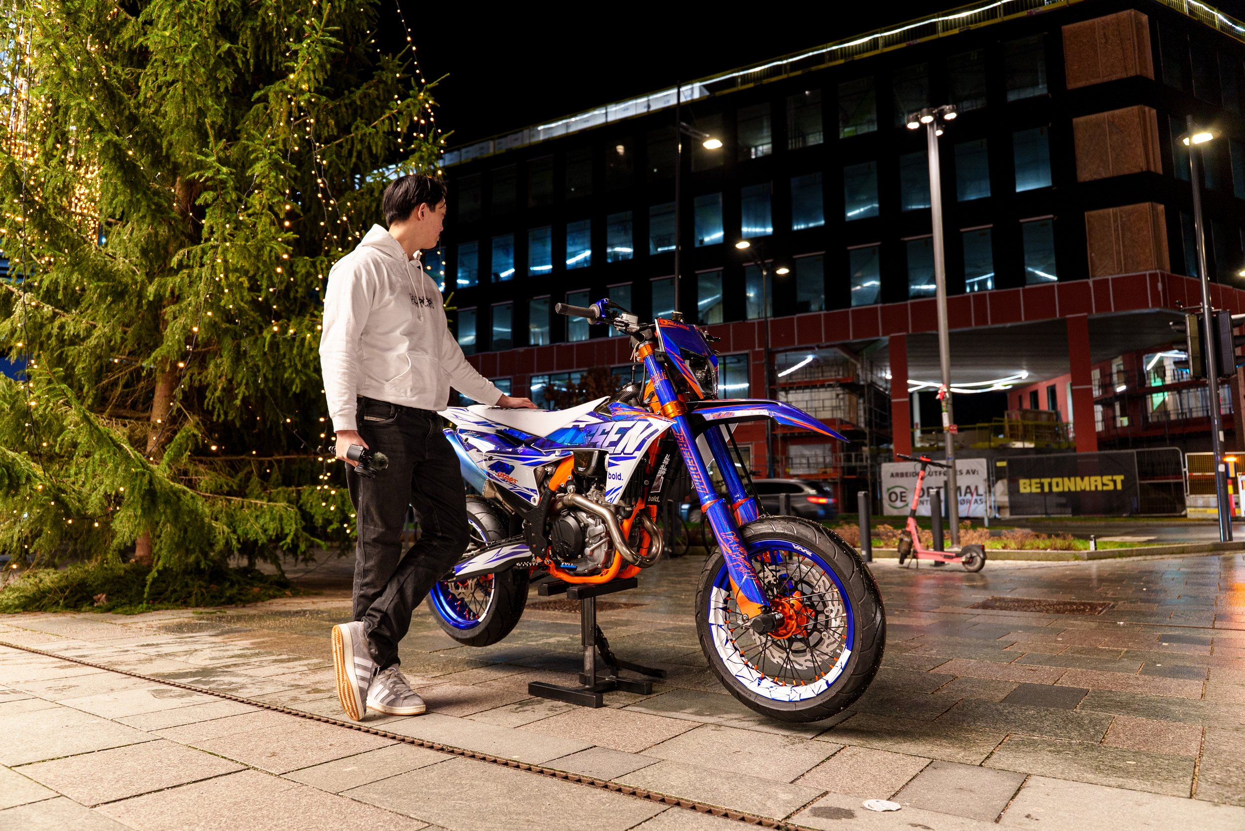 En person står ved en motocross sykkel foran et juletre med lys utenfor en bygning om kvelden.