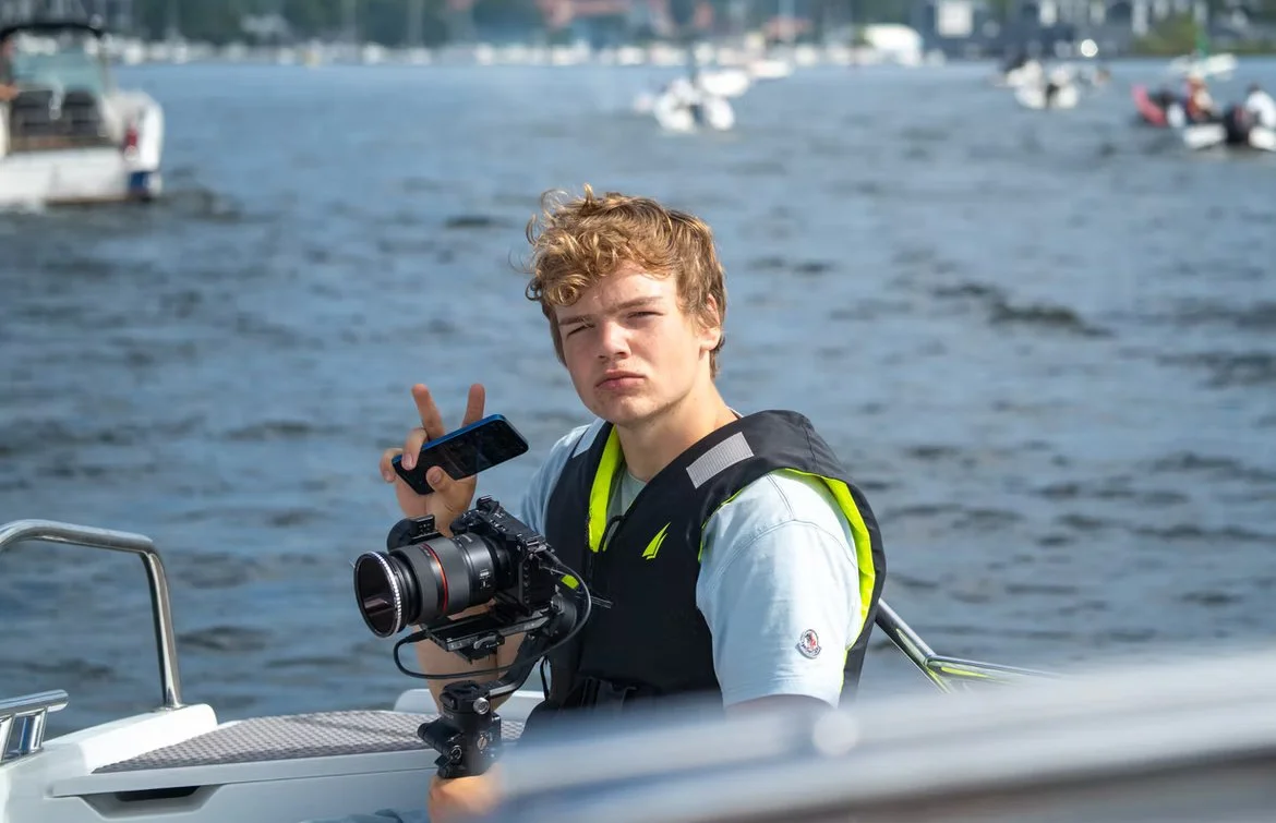 Teenage boy with curly blonde hair on boat holding a camera with a stabilization rig, making a peace sign with his right hand, on a waterway with boats in the background.