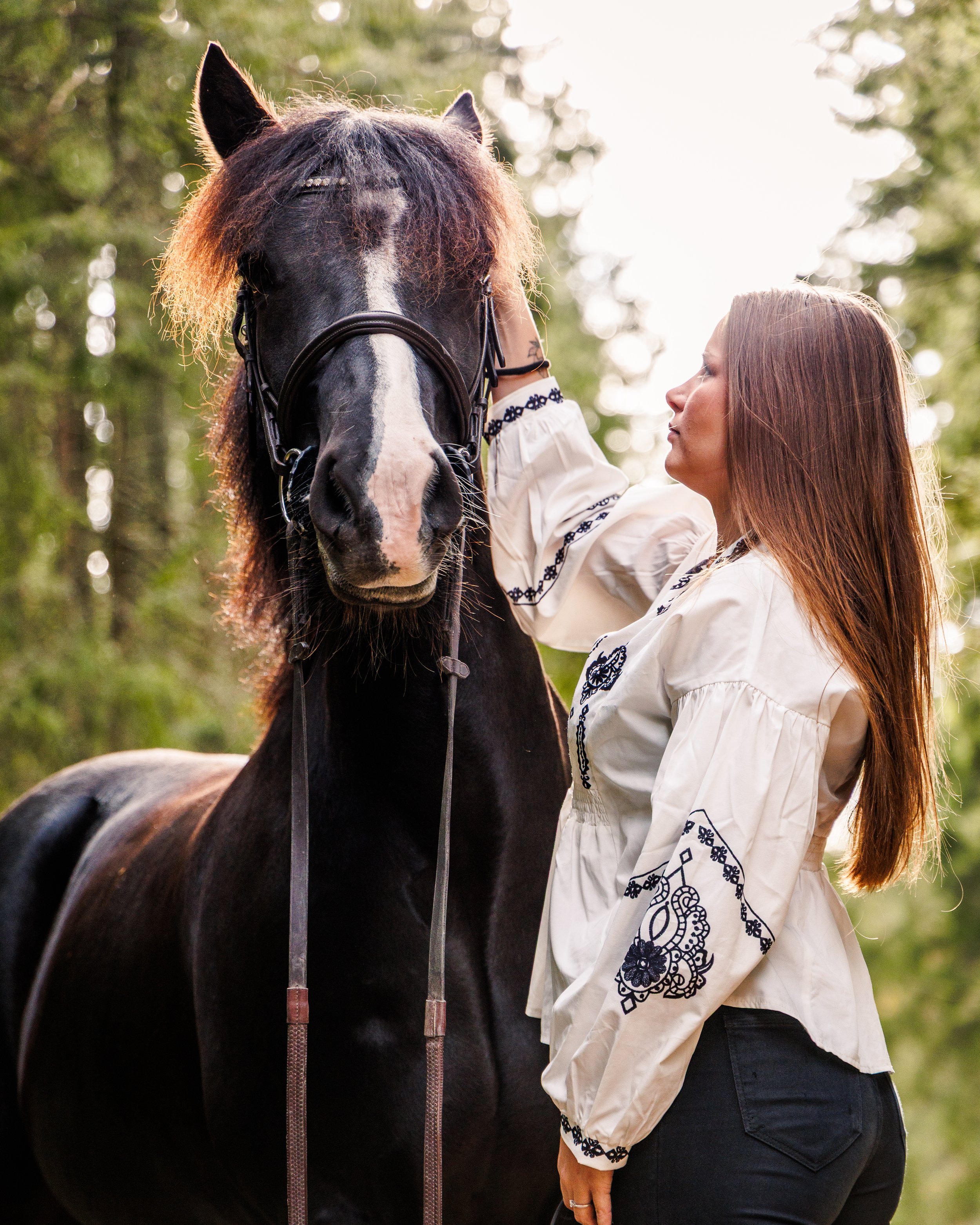 En kvinne med langt brunt hår og en hvit bluse med blå blomstermønster står ved en svart hest med en hvit stripe i ansiktet, i en skog med grønne trær.