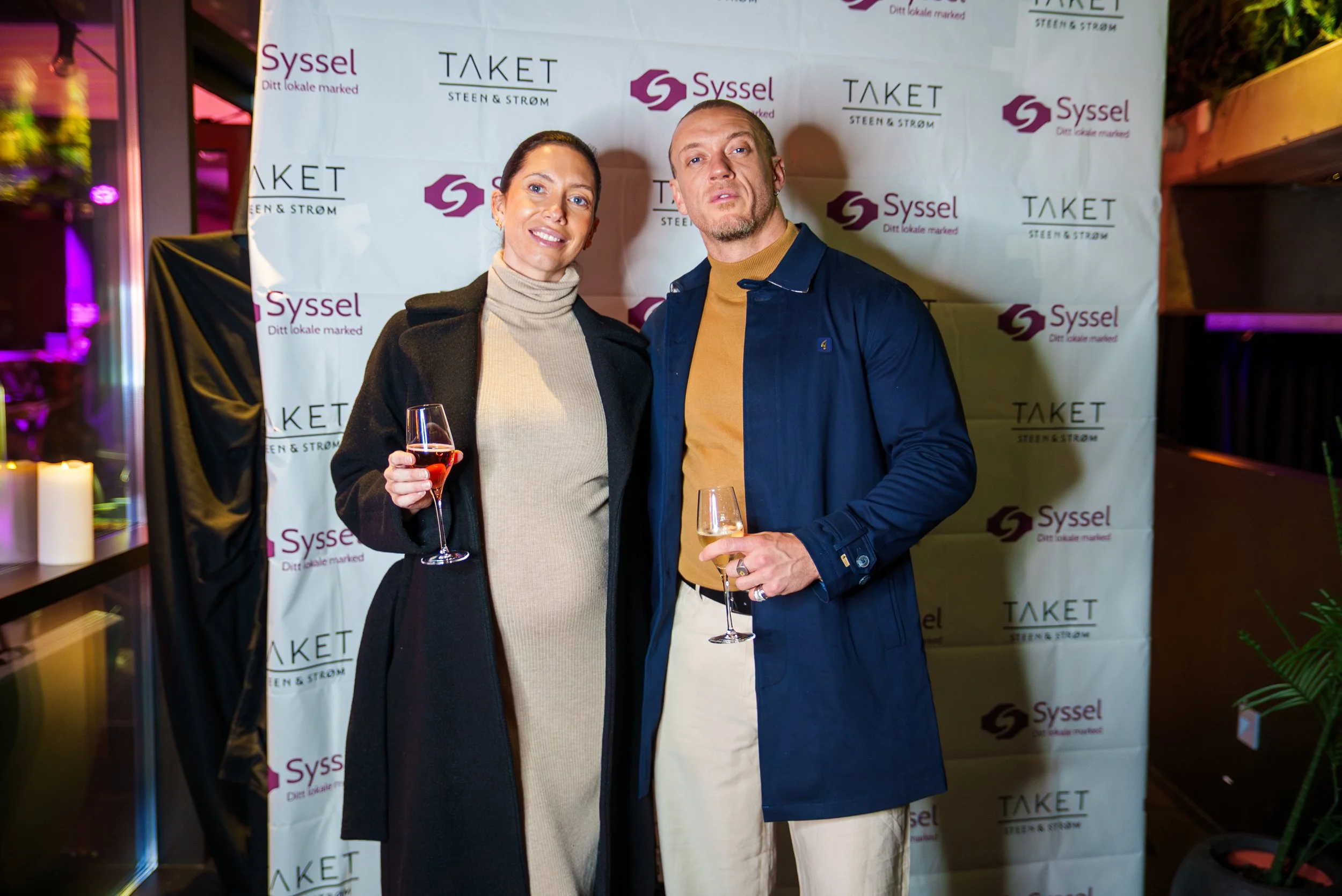 To personer som står sammen med drinker i hånden foran en bakgrunn med logoene '-Syssel', 'Taket', og 'Steen & Strøm'.