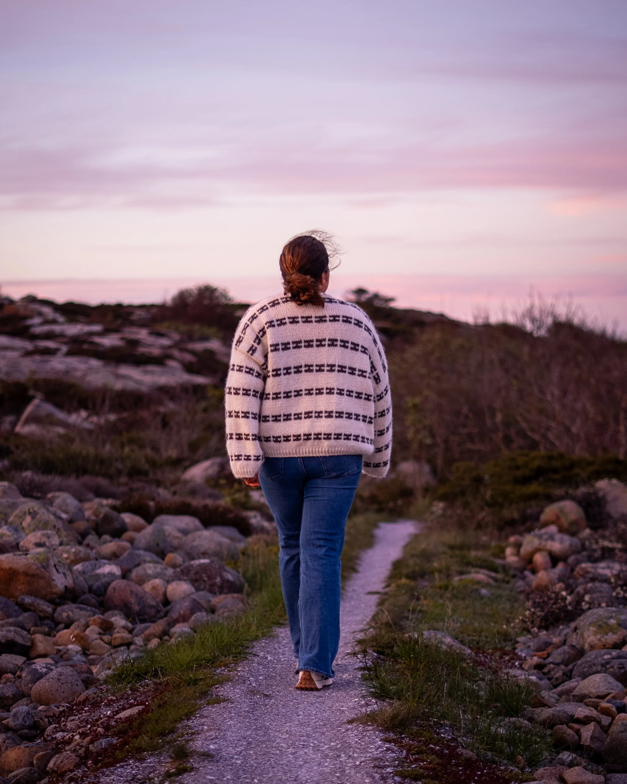 En person går langs en smal sti med steiner og gress, med en vakker solnedgang på himmelen.