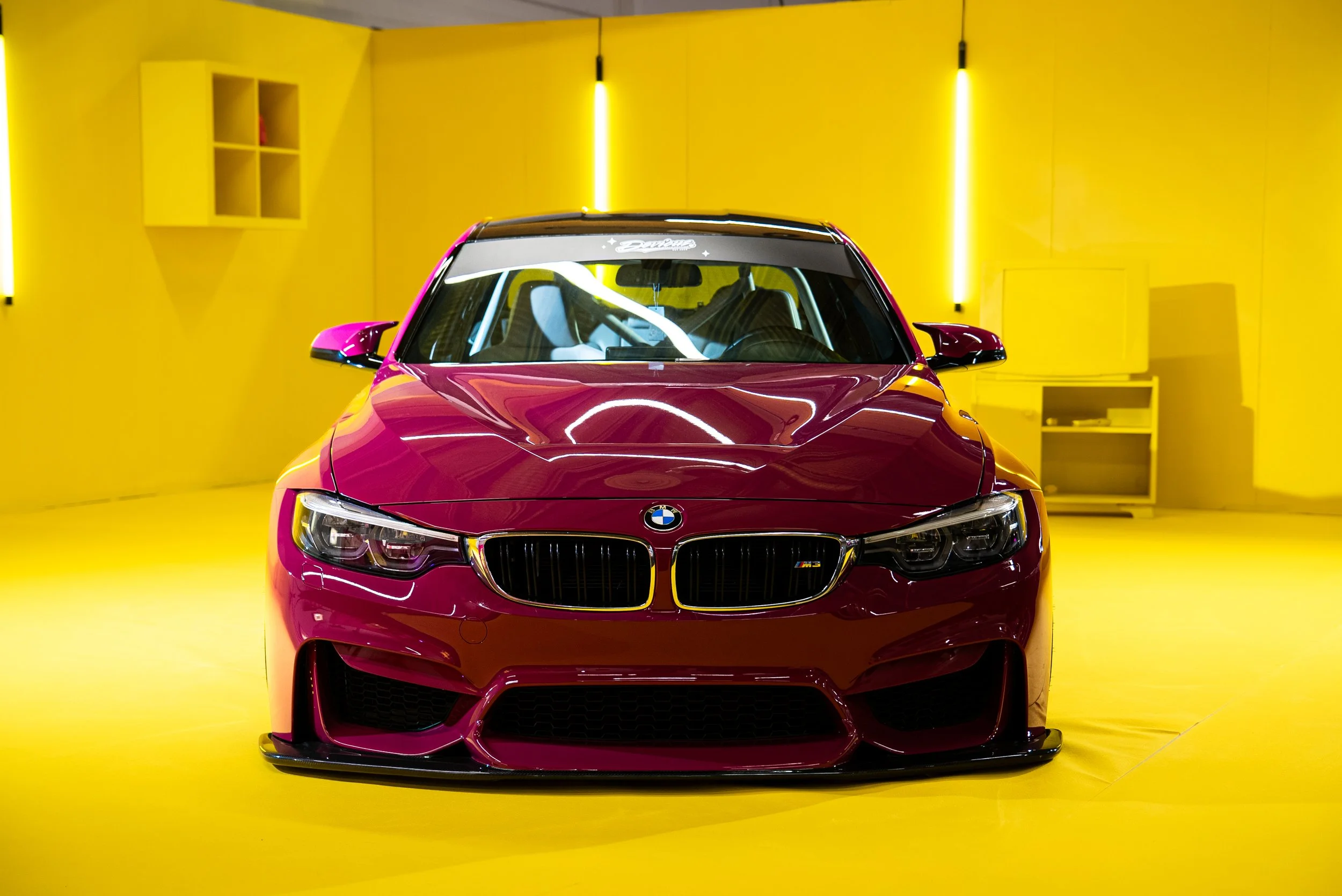 En rød BMW M4 sportsbil plassert på en gul bakgrunn med enkel innredning, opplyst av flere lyskilder.