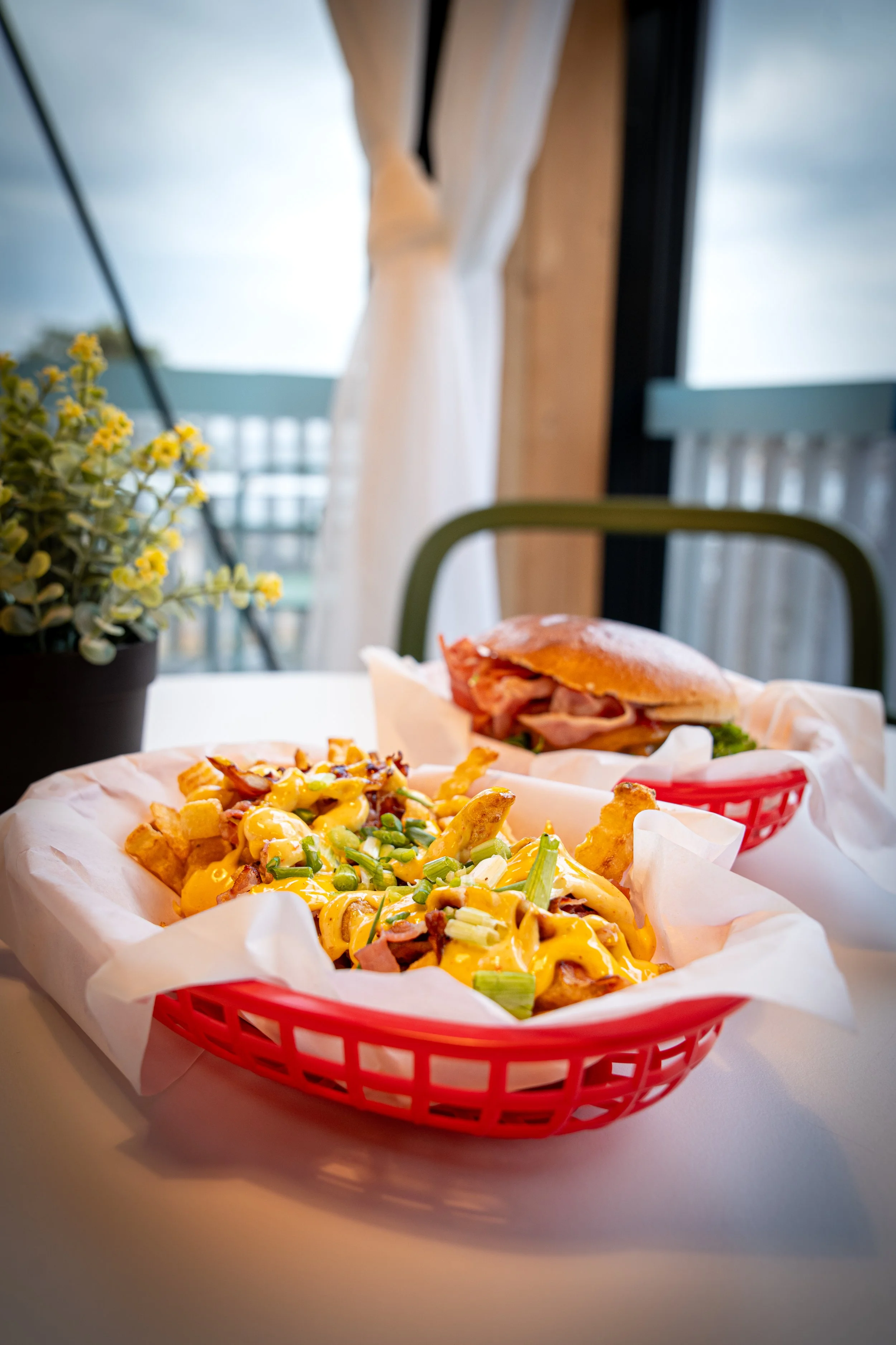 En pappkopp med pommes frites dekket av ost, løk og grønne løkstenger, og en sandwich med kjøttpålegg og salat i bakgrunnen.