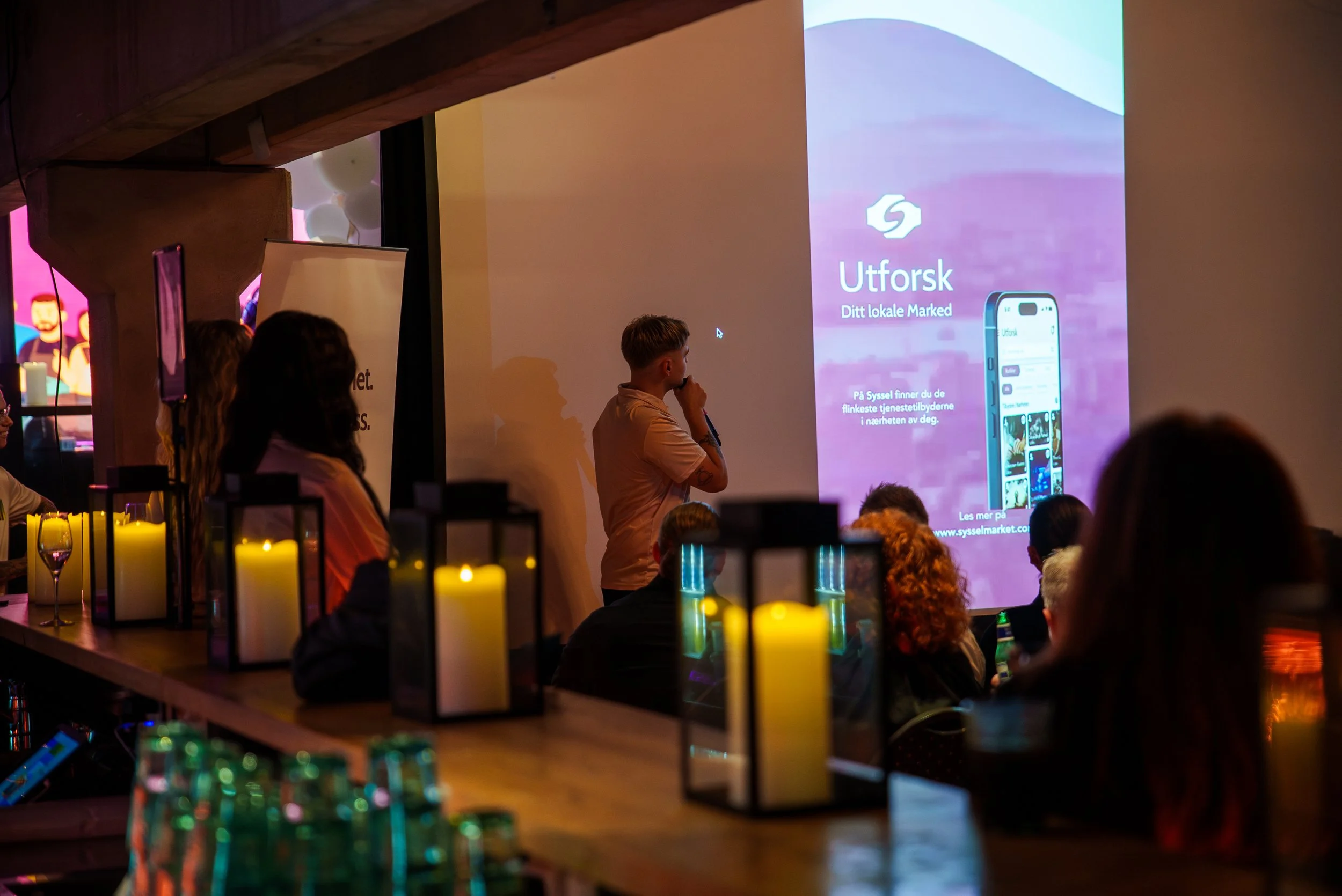En person holder en mikrofon foran en stor skjerm som viser en presentasjon med tekster og en telefon. Publikum sitter i et rom med dekorerte lyslykt på en lang bar i forgrunnen.