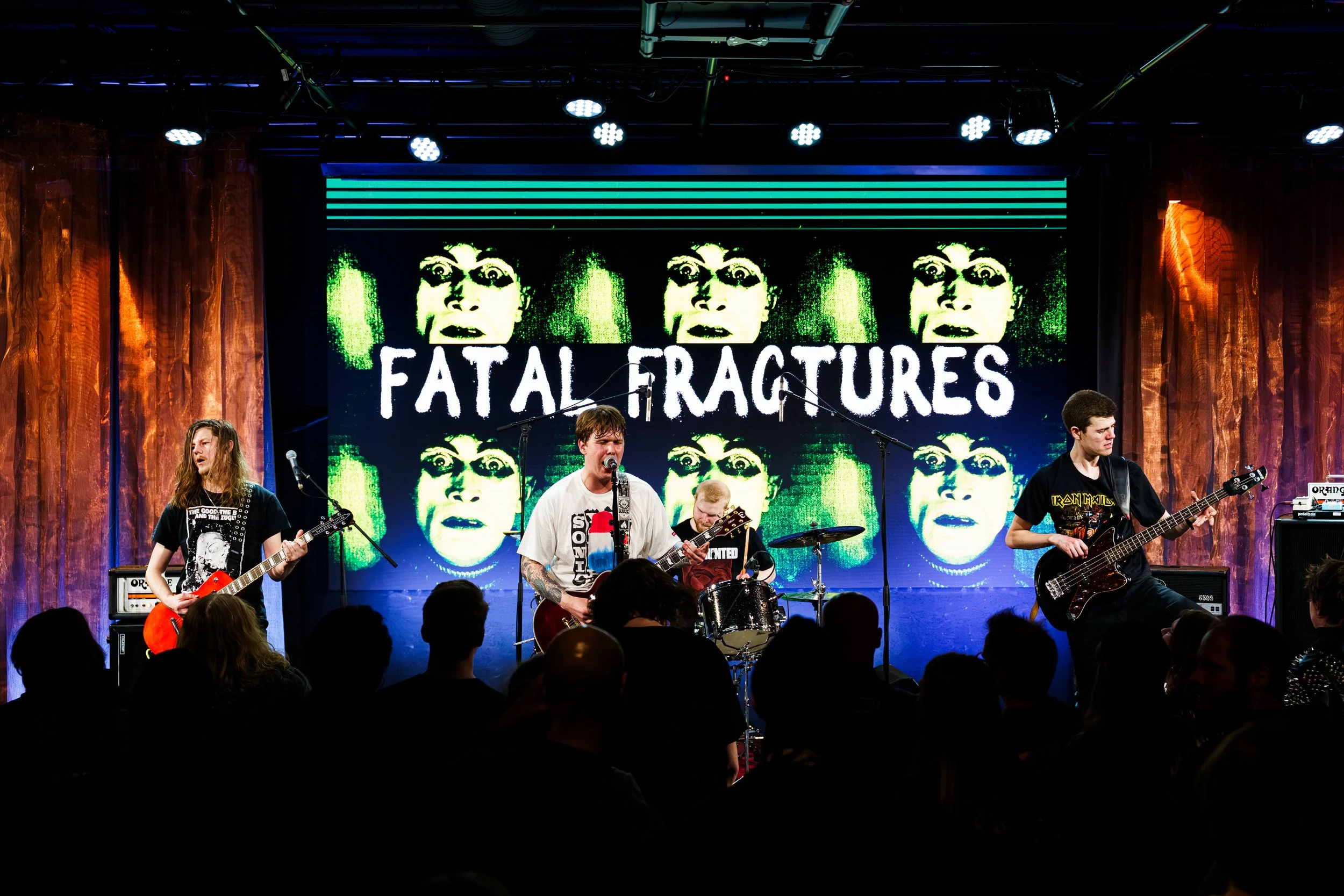 Et band spiller på scenen under en konsert med et digitalt bakgrunnsdisplay som viser bildet av en persons ansikt og teksten "FATAL FRACTURES".