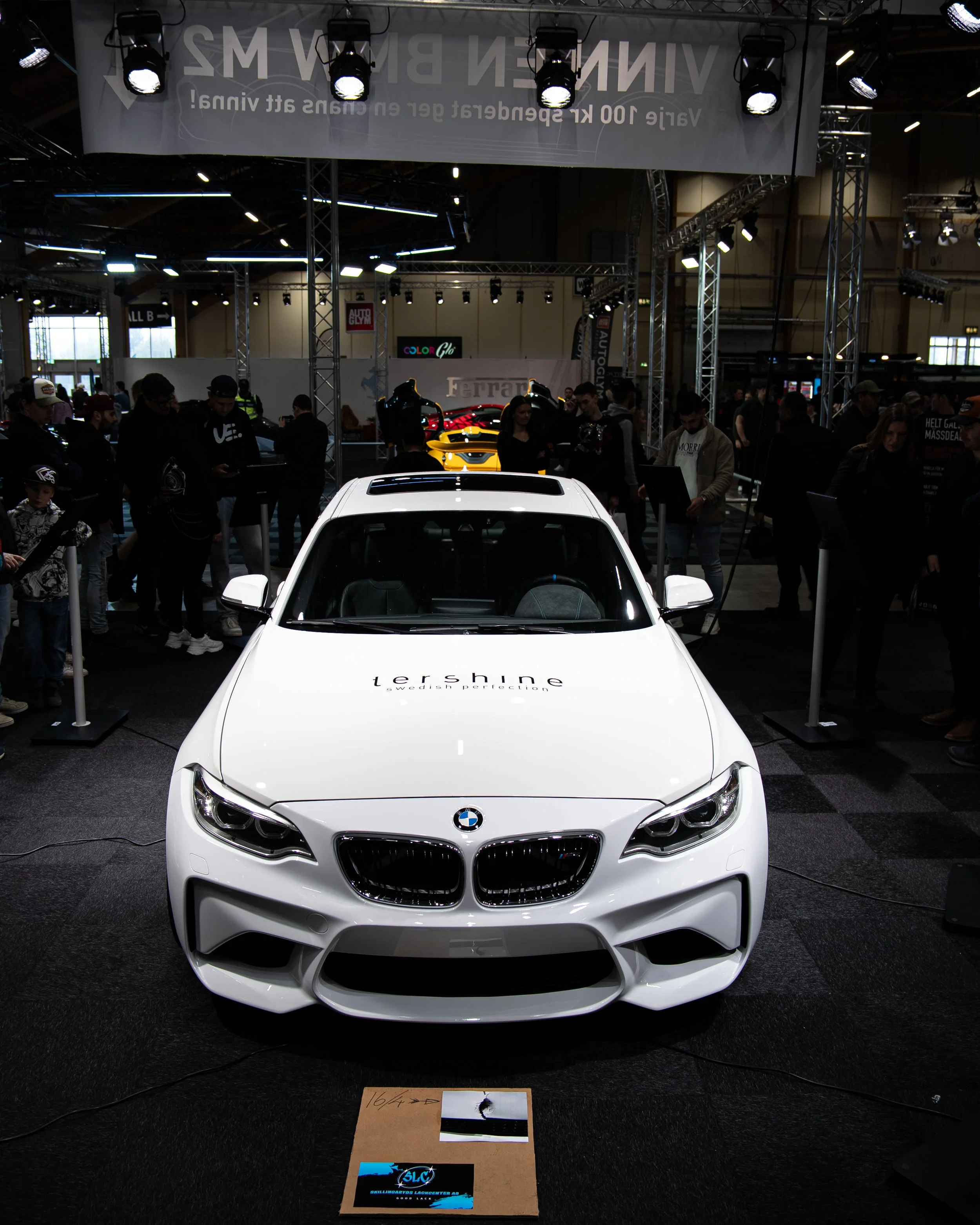 En hvit BMW- bil utstilt på en messe, omgitt av folk og andre biler, med belysning og skjermer rundt.