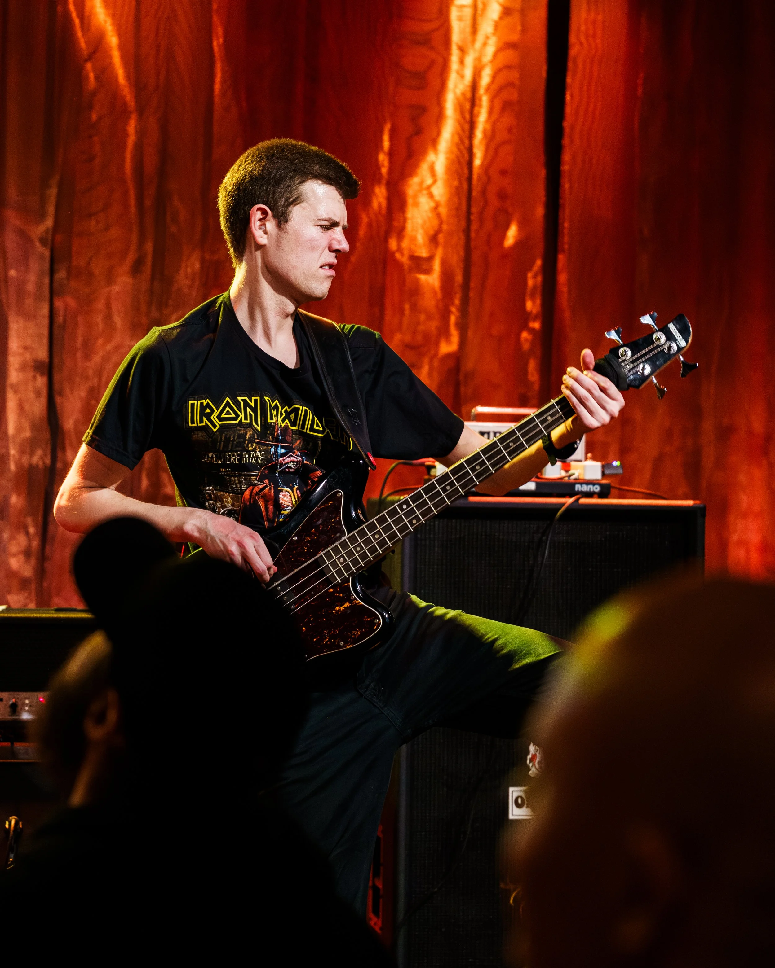 En mann spiller bassgitar på scenen med lysebrunt treverk som bakgrunn.