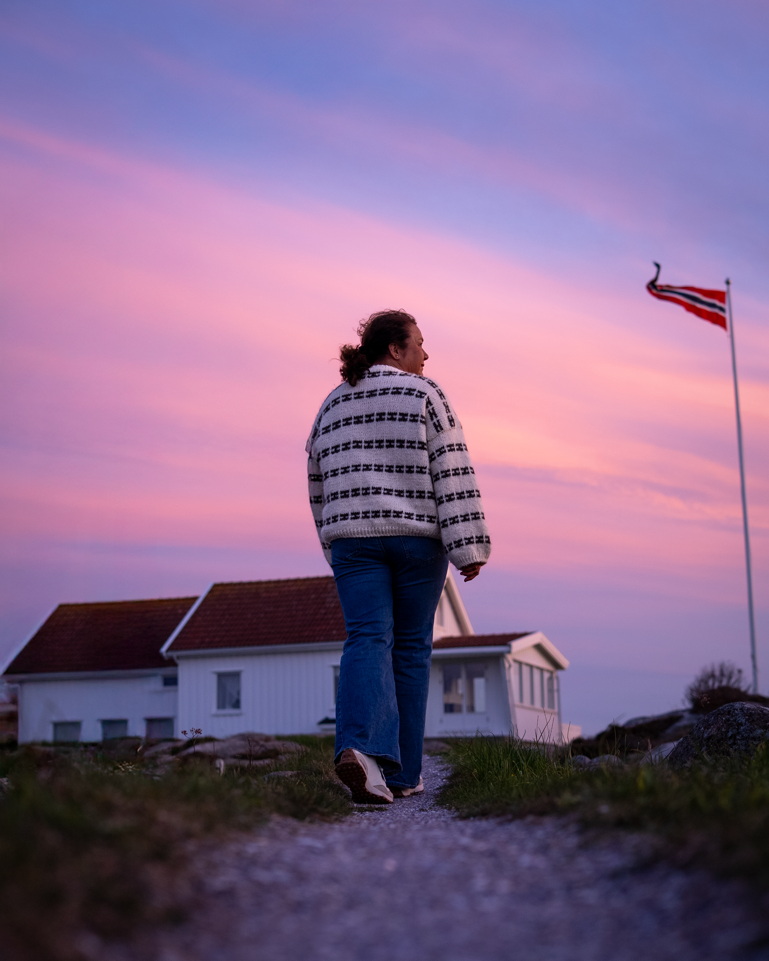 En kvinne går på en grusvei mot en hvit hytte under en fargerik solnedgang, med en norsk flaggstang i bakgrunnen.