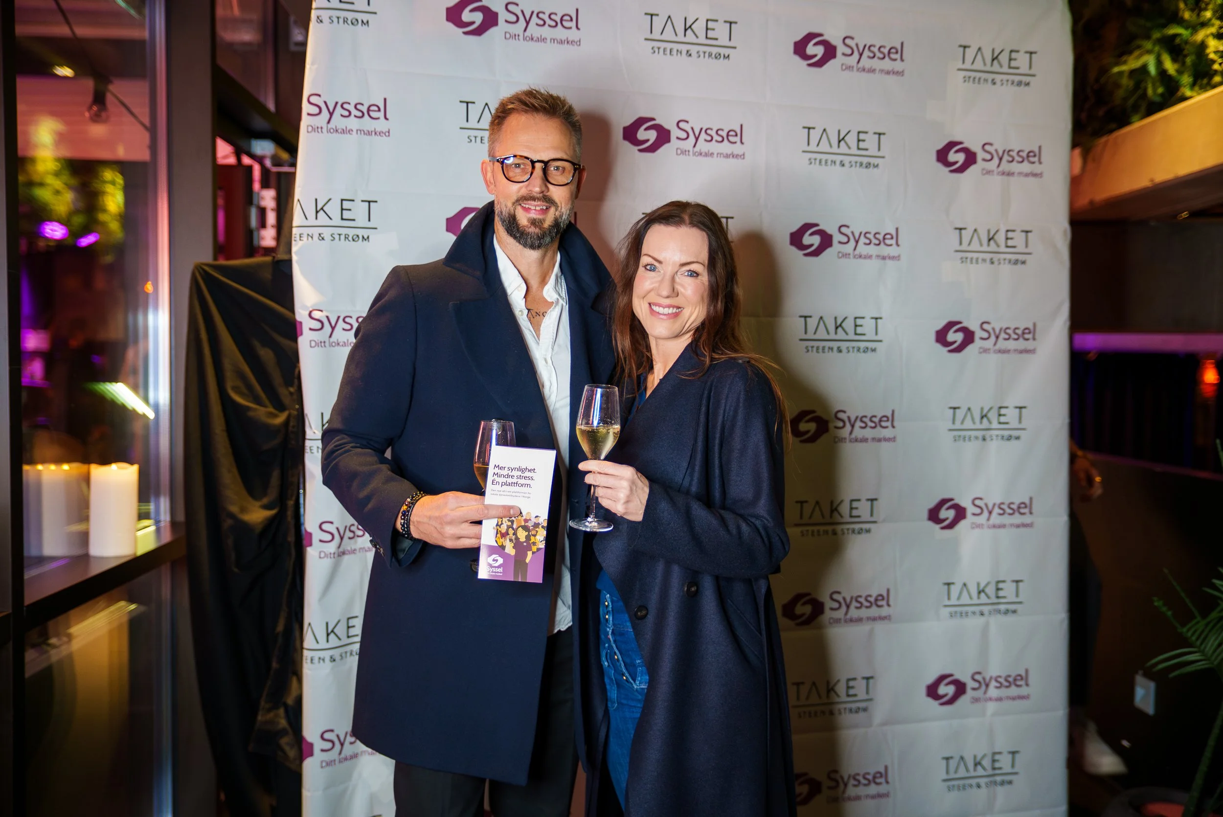 To personer, en mann og en kvinne, smiler og holder glass med boblevann på en fest, foran en backdrop med logoene til Syssel og Taket.