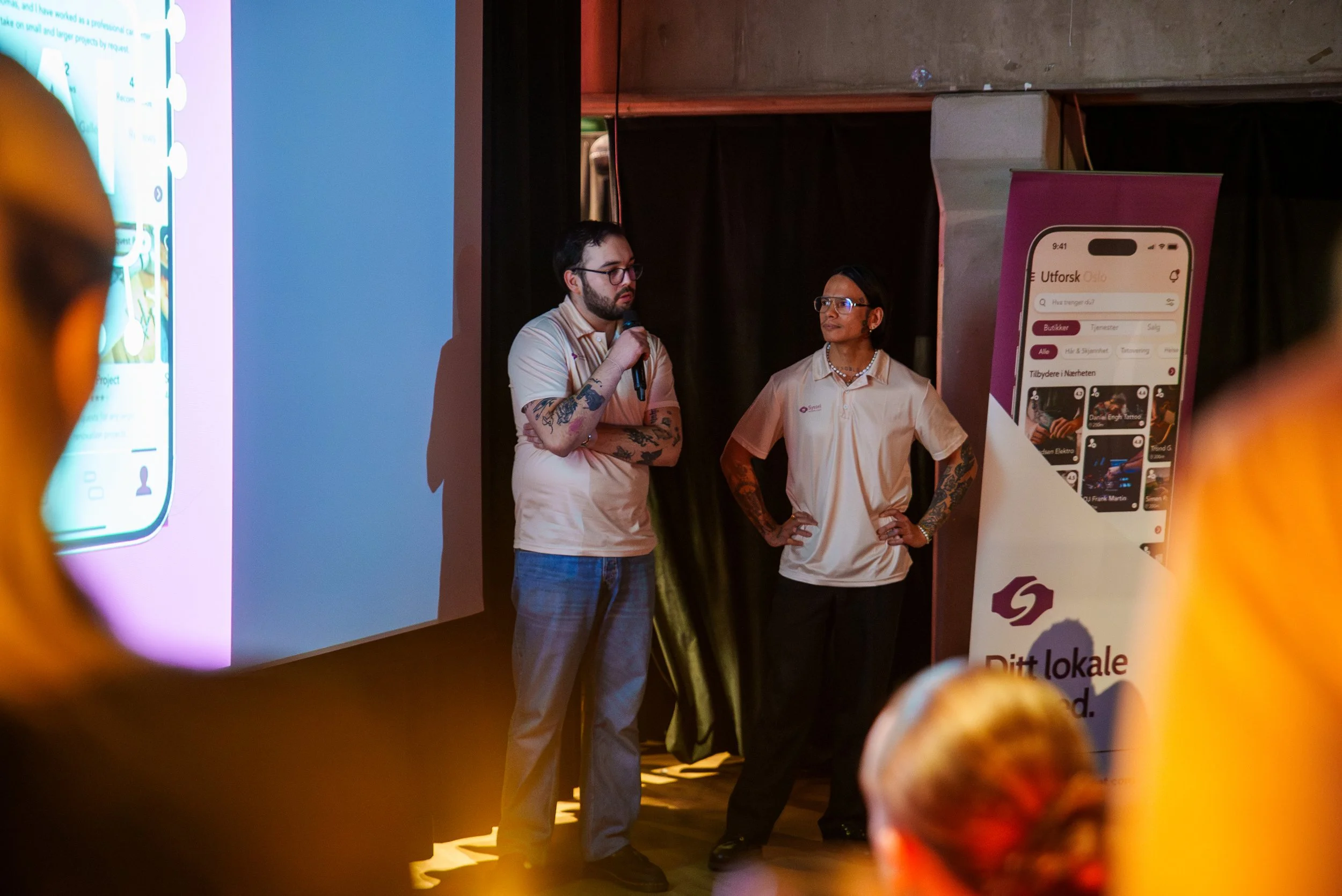 To personer snakker foran en stor skjerm og en banner med en mobilapp, hvor den ene personen holder mikrofon og den andre står med hendene på hoftene. Publikum er synlig i forgrunnen.