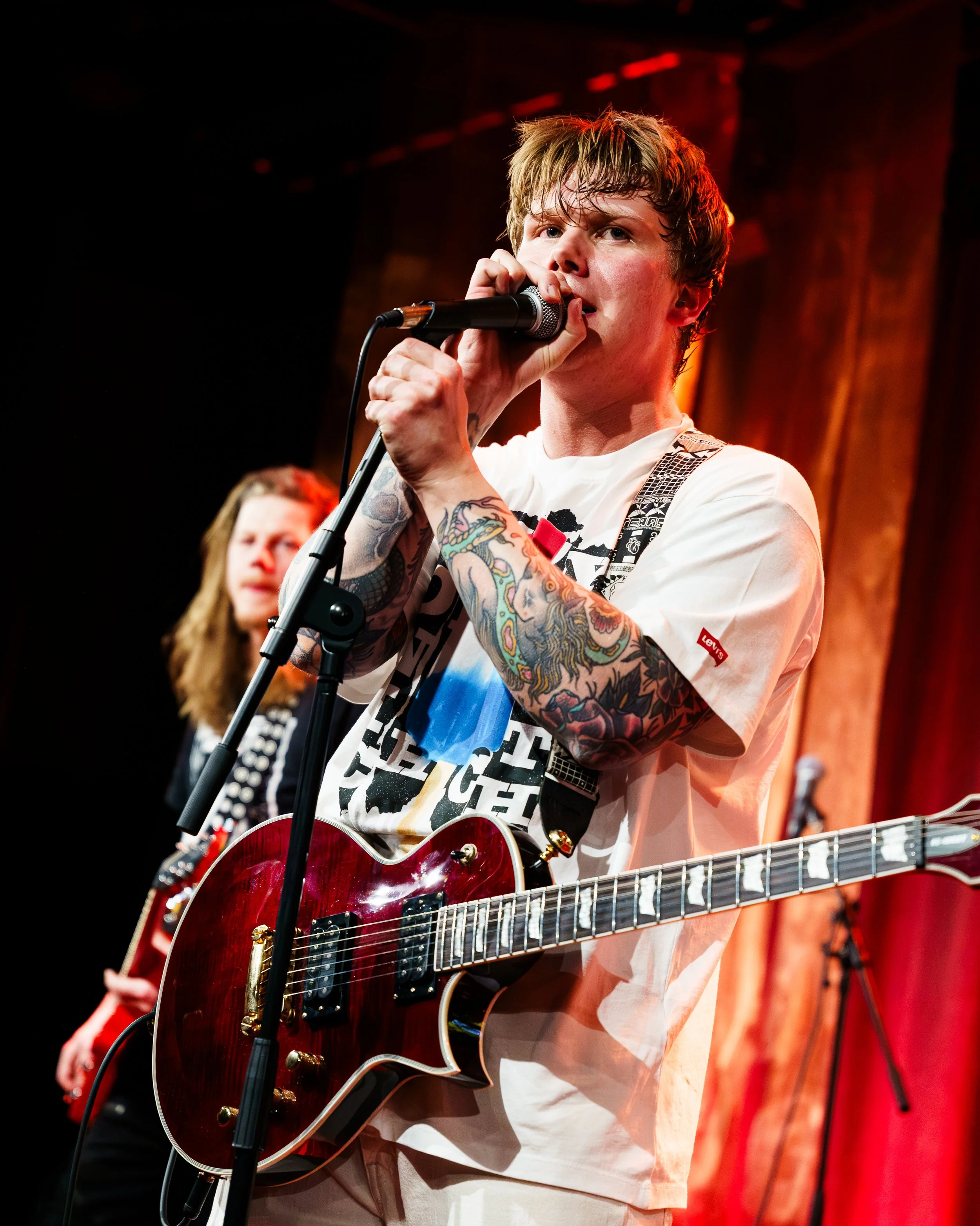 En ung mann med tatoveringer som spiller inn musikkinstrumenter, synger i mikrofon, og står på en scene med rød bakgrunn.