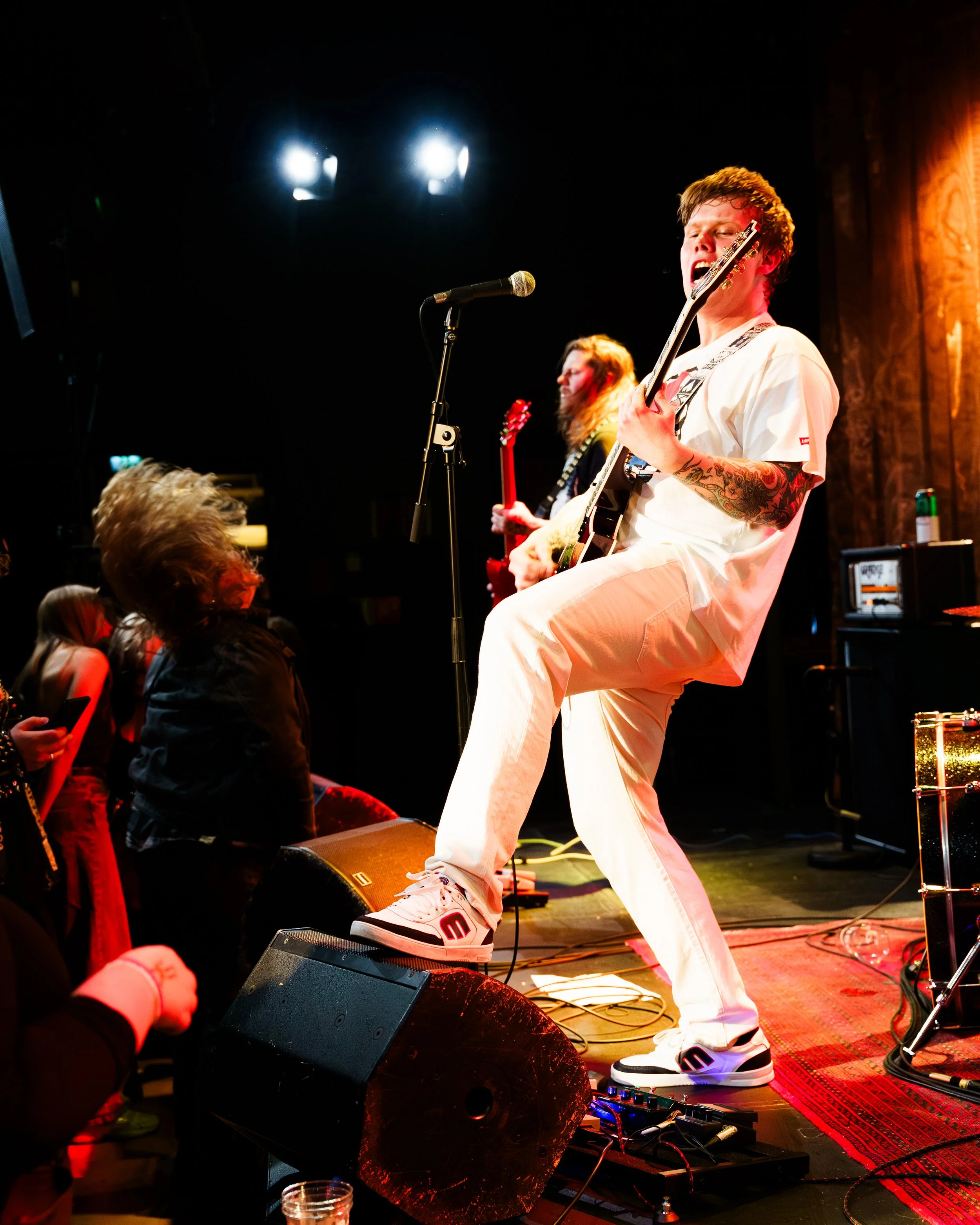 En ung mann spiller gitar og synger på en scene, med en annen musiker i bakgrunnen, under en konsert.