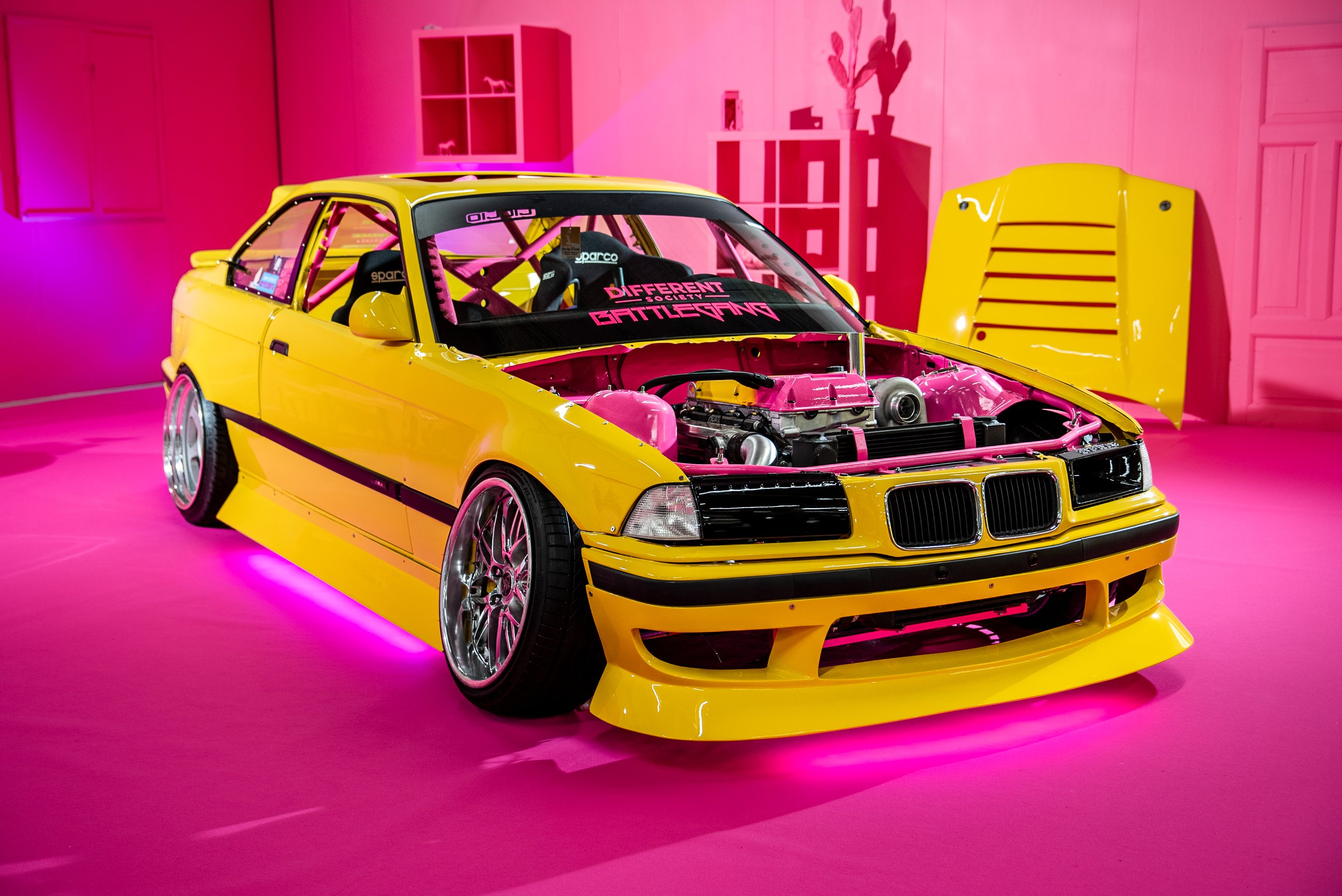 Fremvisning av en delvis stripet gul racerbilm med åpent panser, rød bakgrunn og rosa gulv, med ulike tilbehør og utsmykking. 