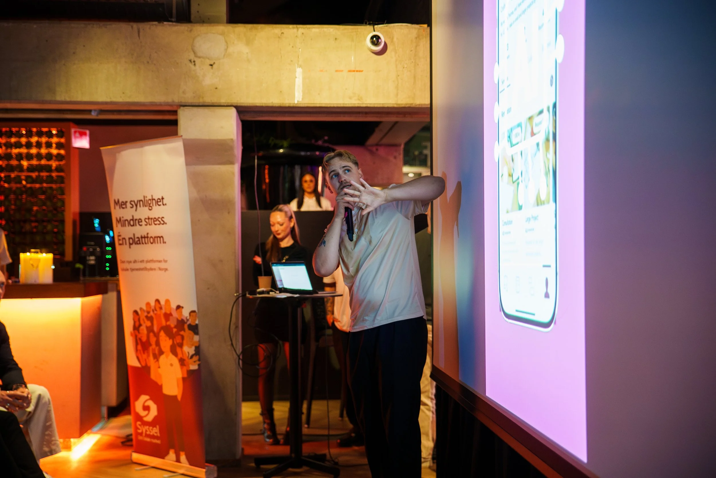 En mann holder en mikrofon og peker mot en stor skjerm med en mobilapp på. Det er et arrangement med flere deltakere og en banner med tekst på venstre side.