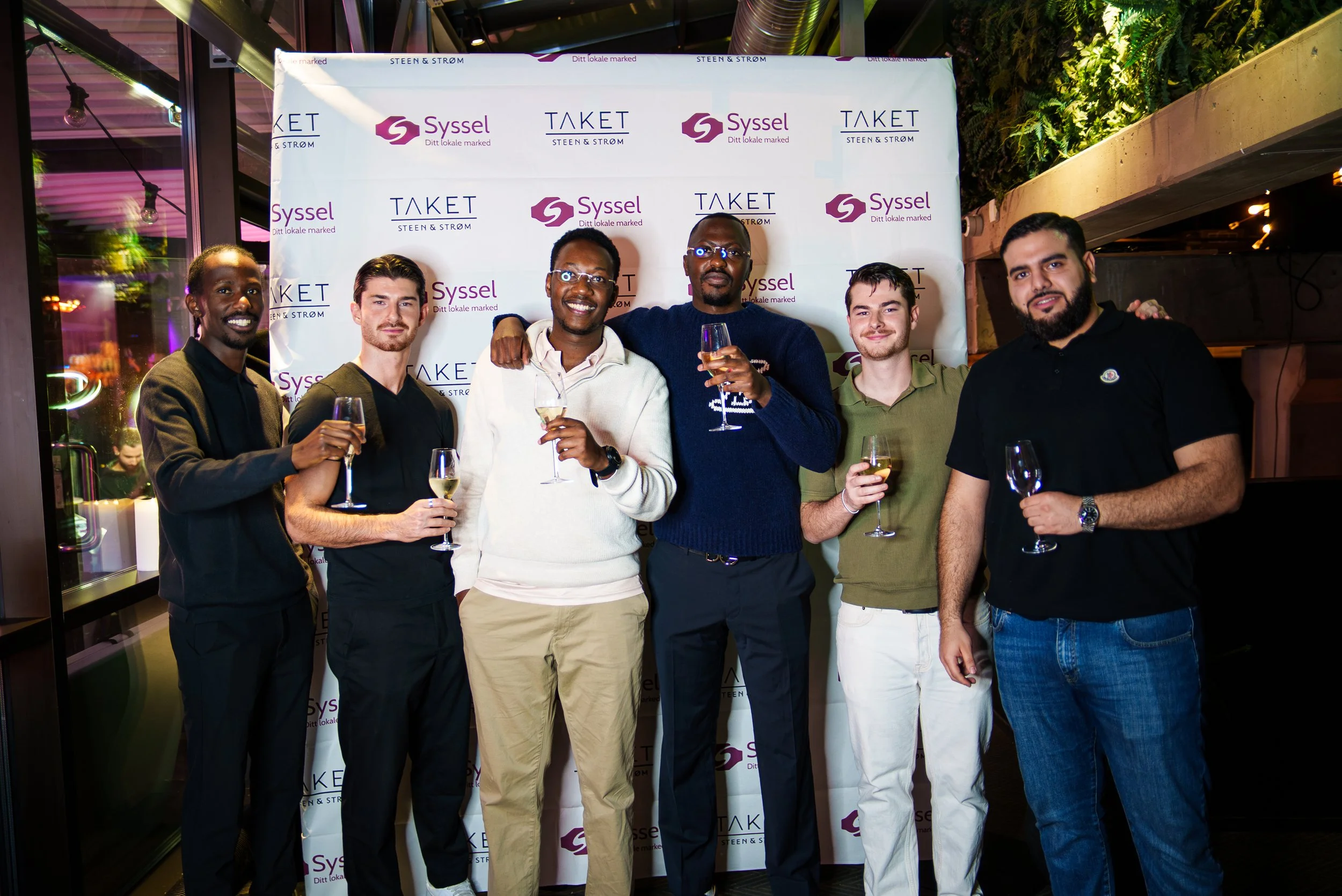 Syv personer står sammen og holder glass med vin, foran en bakgrunn med logoene til Syssel og Taket, i en innendørs setting med dekorasjoner og belysning.