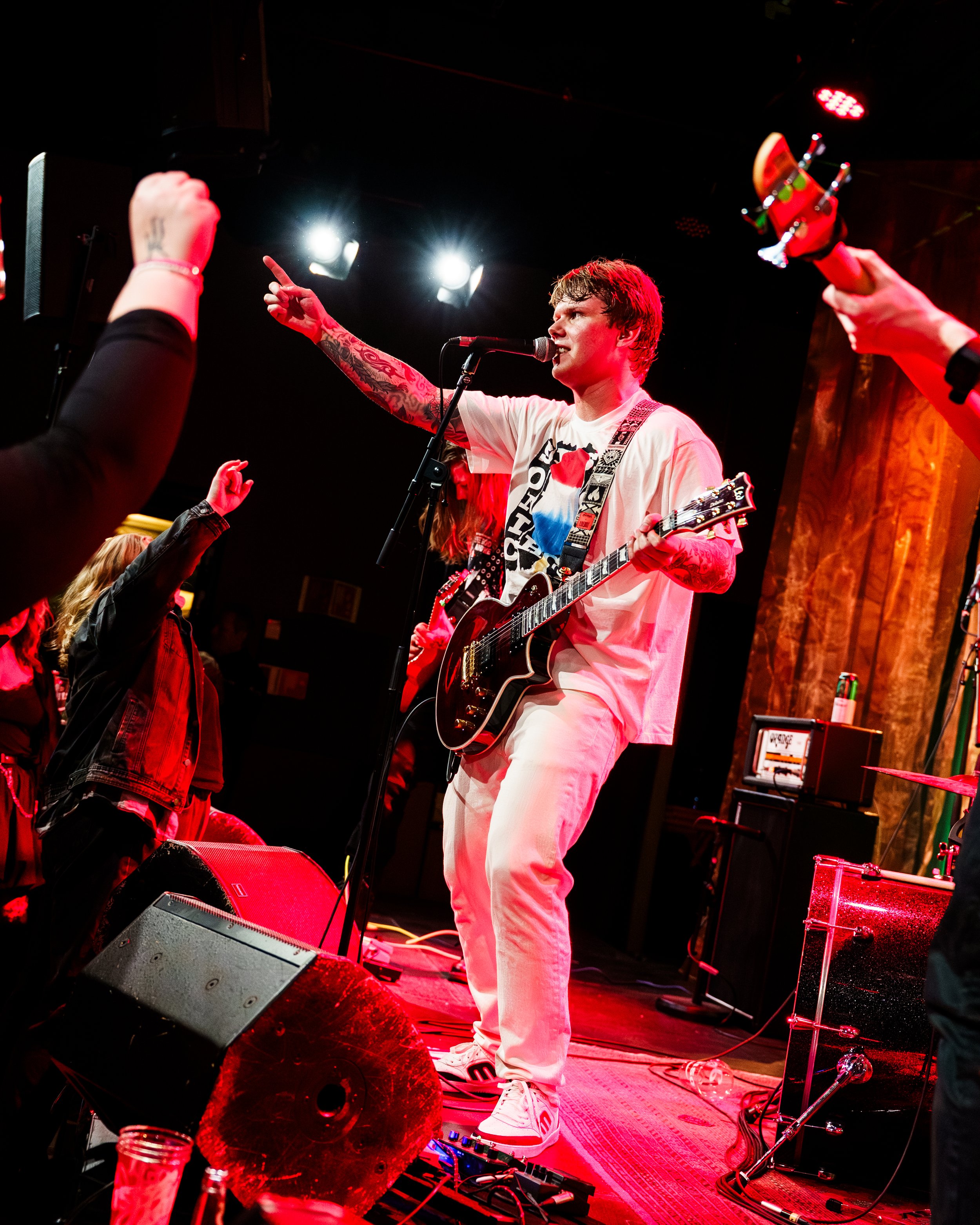 En mann spiller elektrisk gitar og synger inn mikrofon på en konsertscene, omgitt av folk som danser og heier.