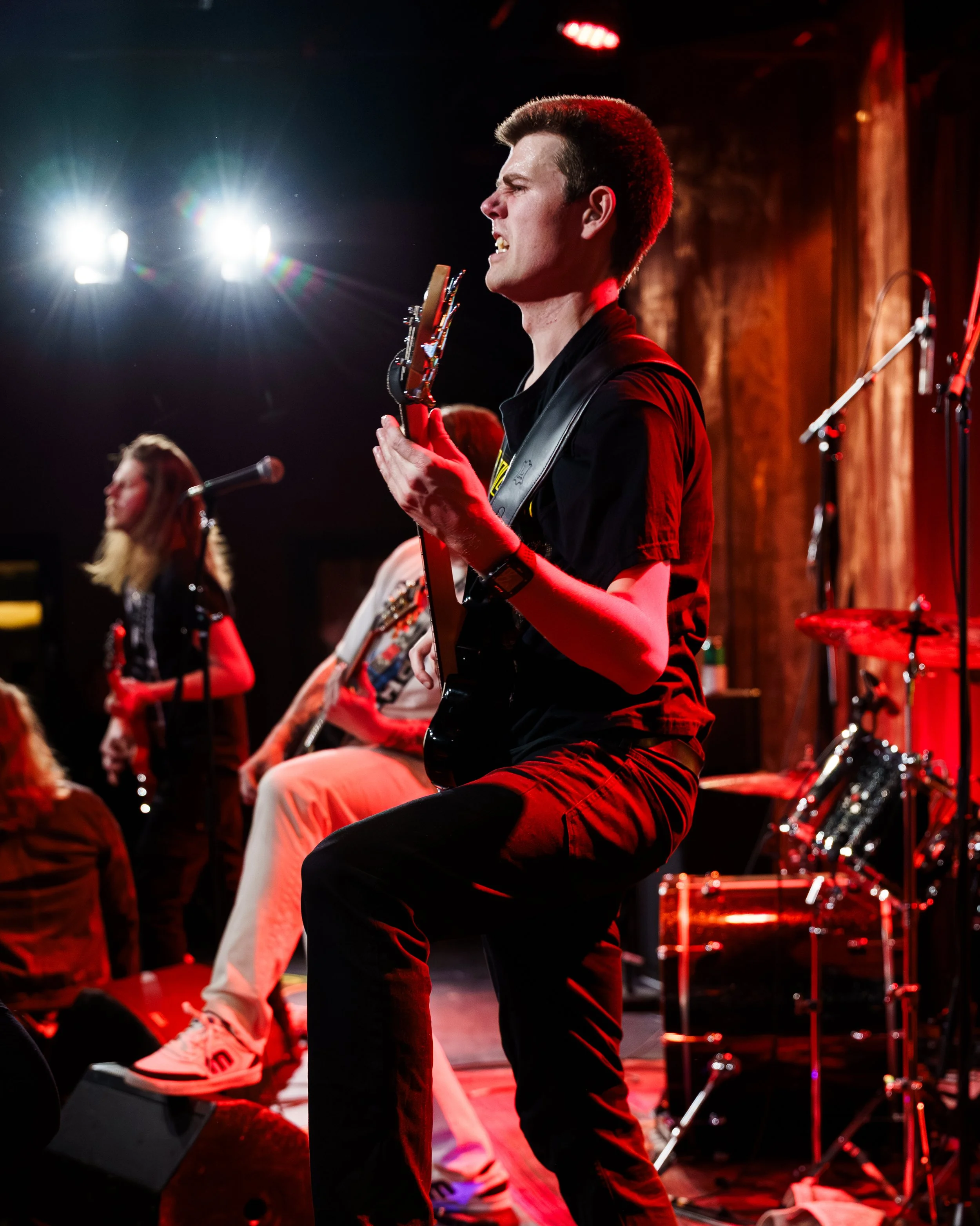 En mann spiller elektrisk gitar på en scene med rødt og svart lys, omgitt av andre musikere.