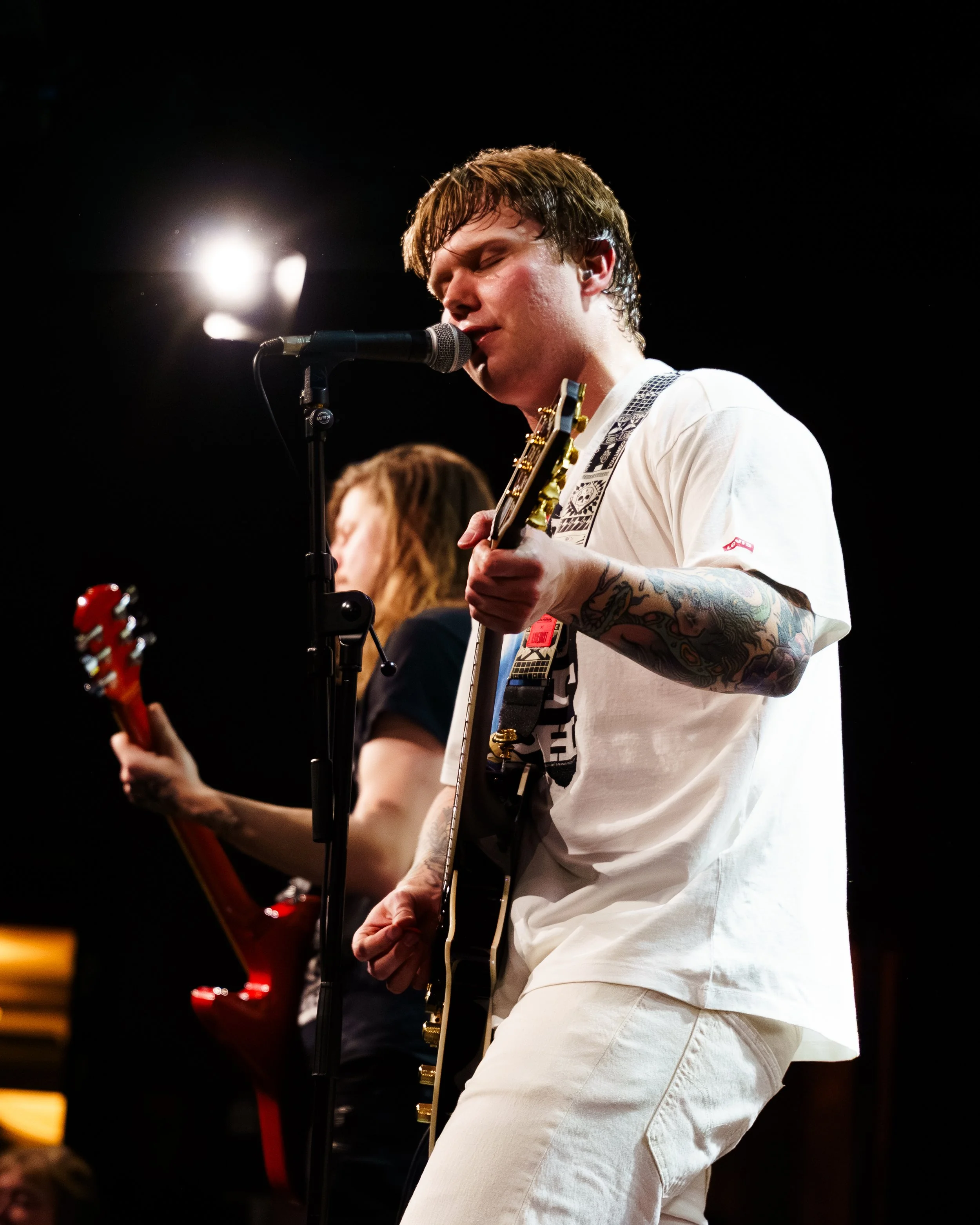 To musikere spiller gitar på scenen, med en av dem som synger inn i mikrofon, under en konsert.