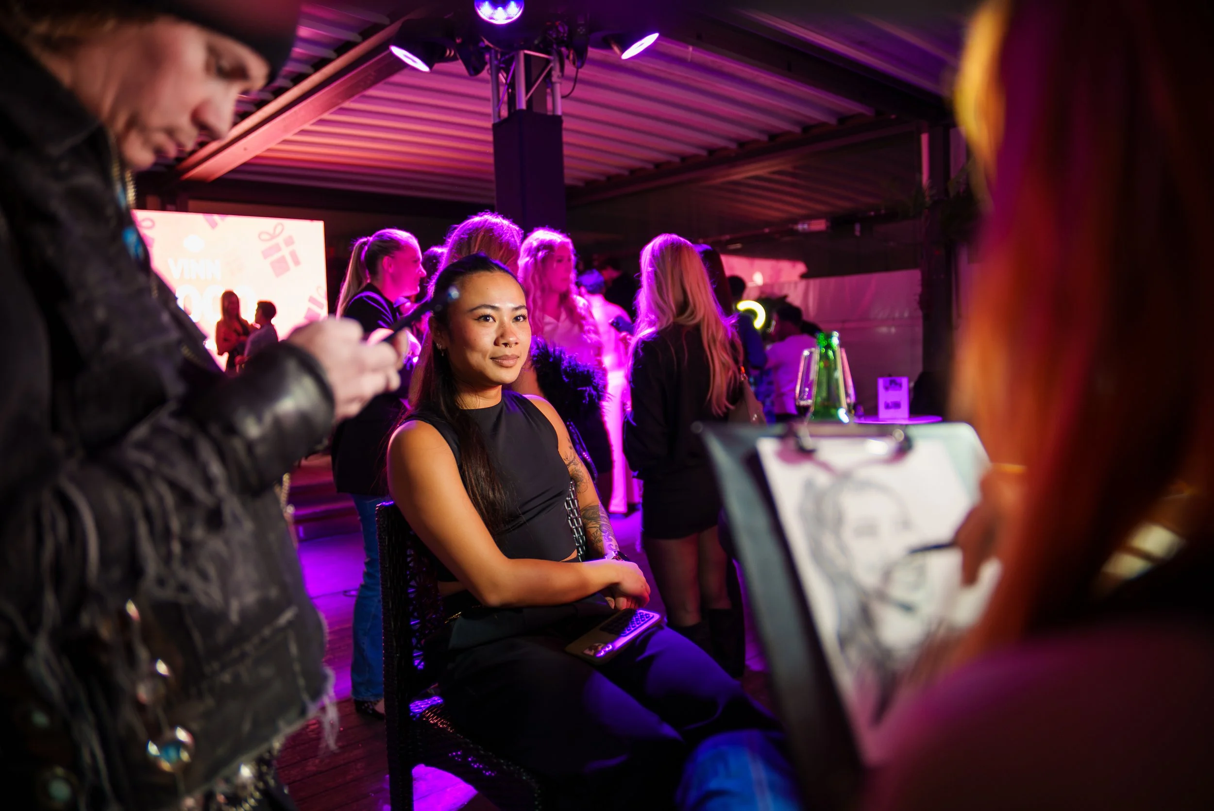 En kvinne med langt mørkt hår og tatoveringer er sittende i en bar eller klubb med rosa og lilla belysning, omgitt av andre mennesker.