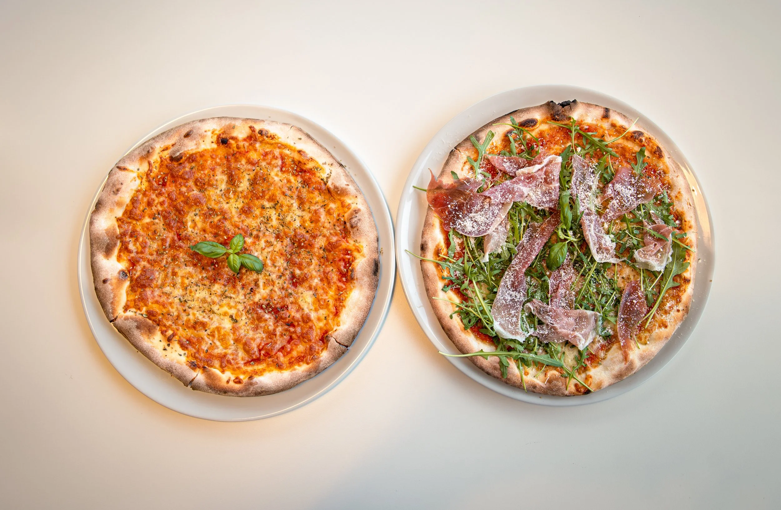 To pizzaer på hvite tallerkener på et lyst bord, en med tomatsaus og ost, den andre med skinke, ruccola og parmesan.