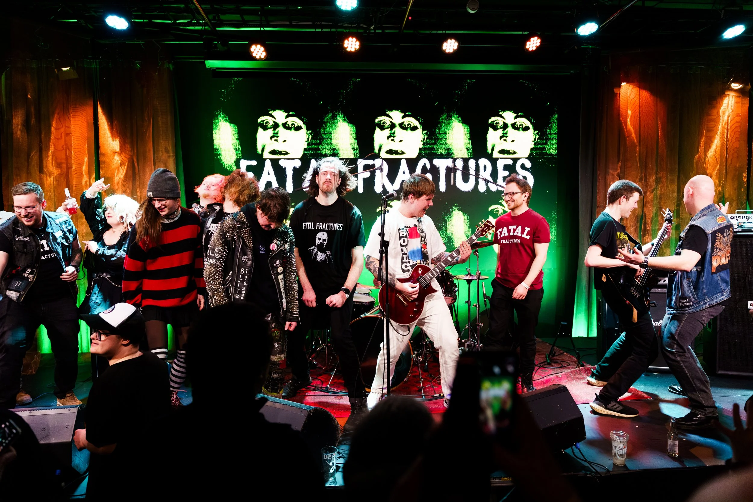 En gruppe musiker på scenen under en konsert med bandet Fatal Fractures, omringet av publikummere, med bandets navn på skjermen bak dem.