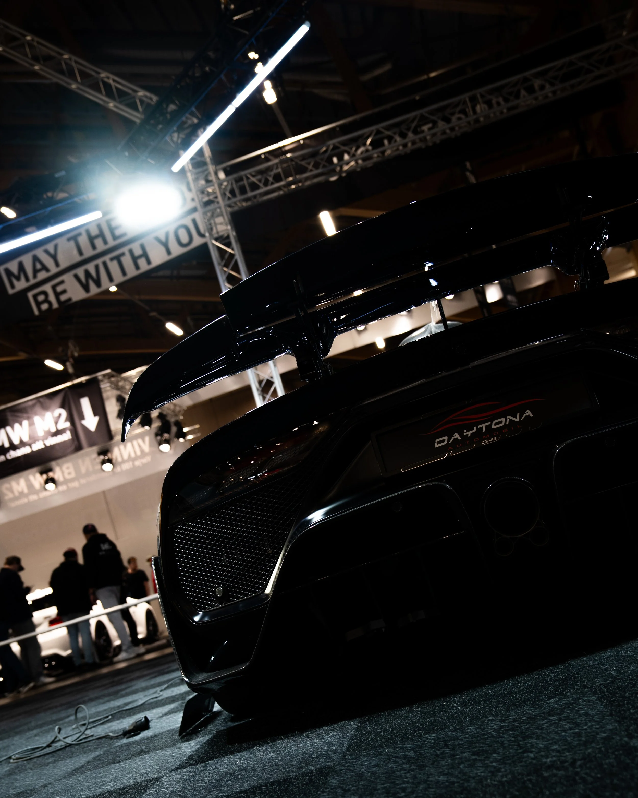 Nærbilde av en svart sportsbil med stor bakspoiler, utstilt på en innendørs utstillingsarena, med mennesker i bakgrunnen.
