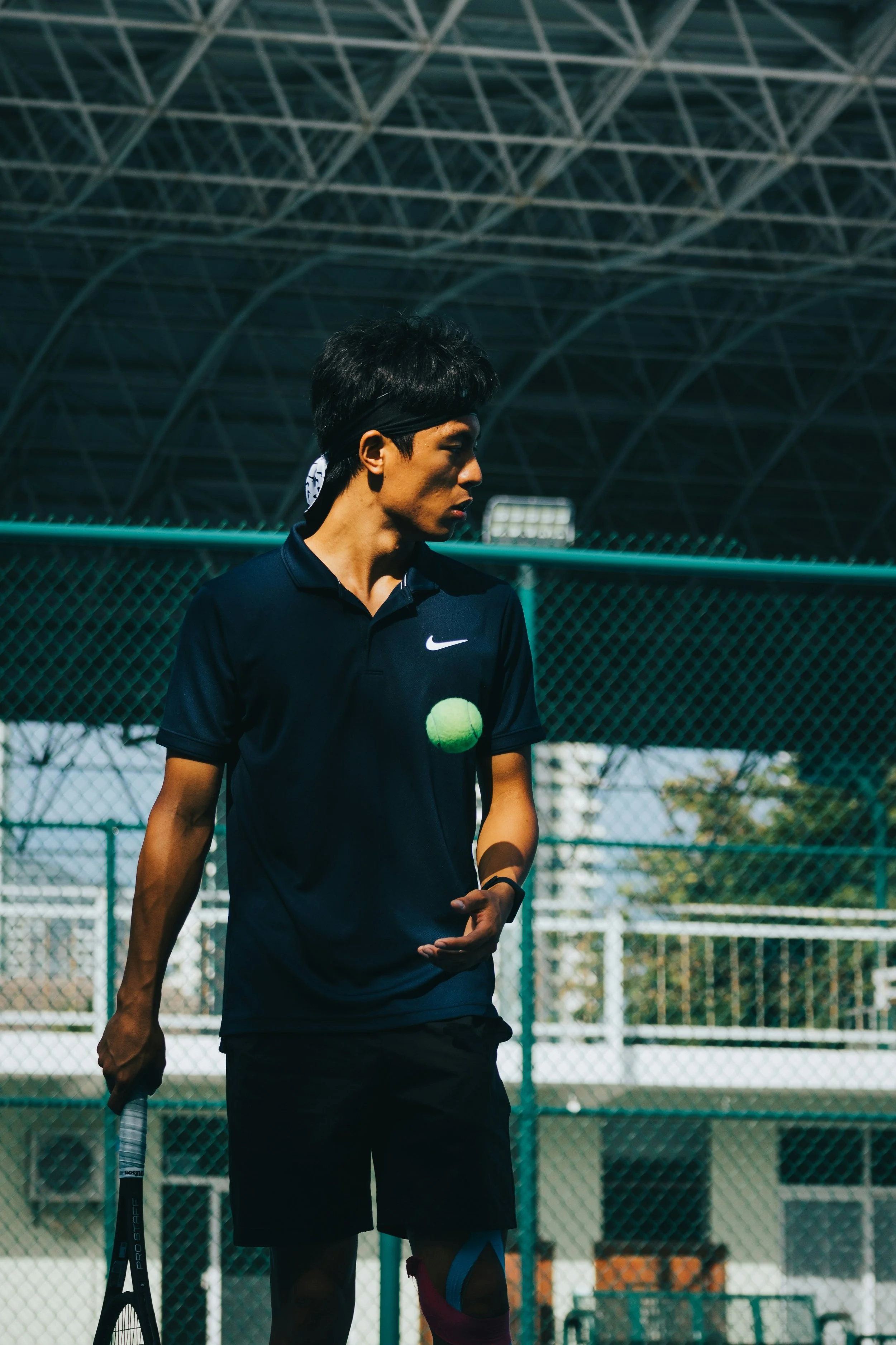 Tennisspeler staat op een tennisbaan met een groene tennisbal en tribune in de achtergrond, draagt een zwart Nike-shirt en heeft een tennisracket in de hand.