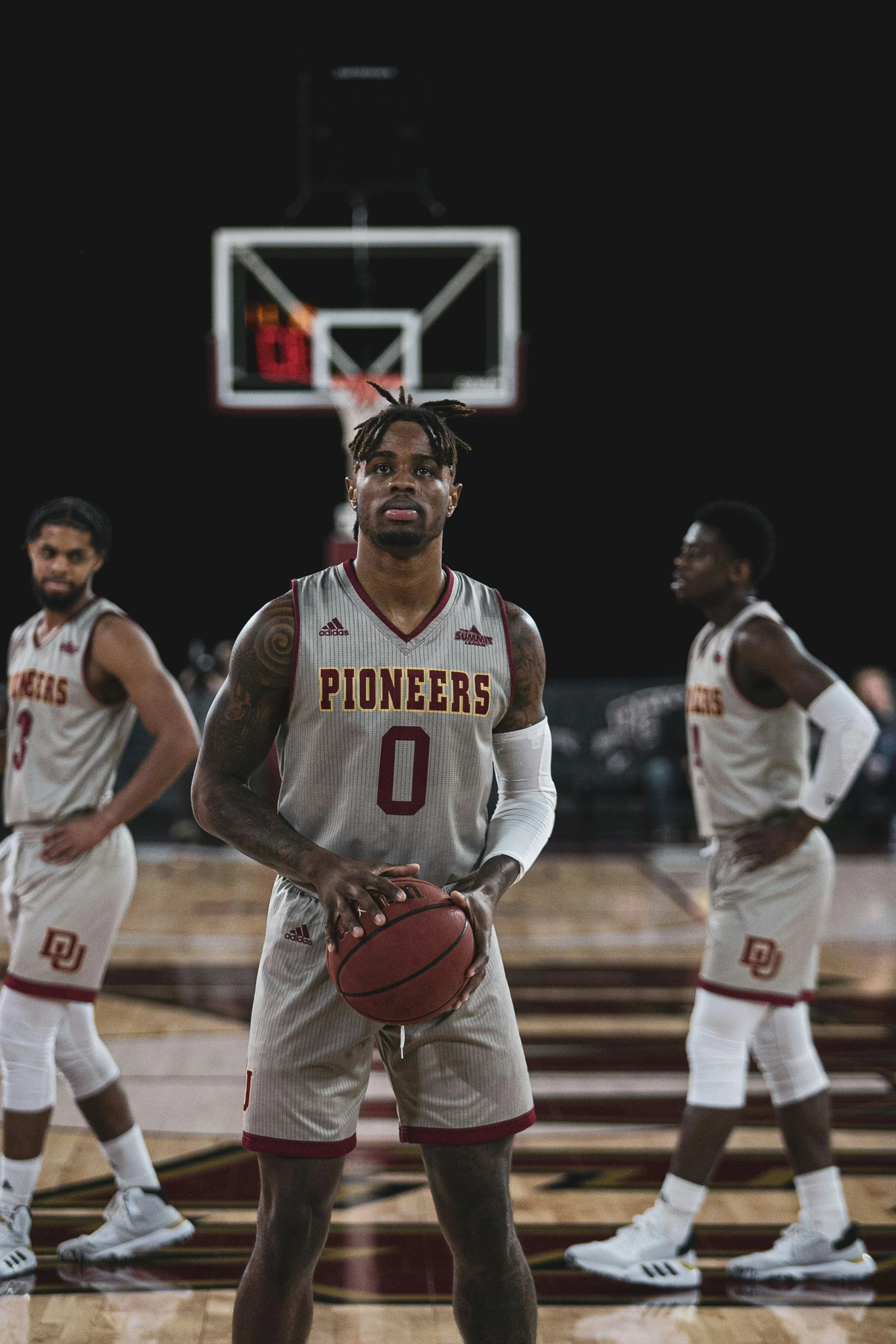 Drie basketbalspelers op een basketbalveld, de speler in het midden met bal en nummer 0 kijkt serieus. De andere twee spelers staan op de achtergrond. Ze dragen witte uniforms met rode letters, het team heet Pioneers, en er is een basketbalring en scoreboard op de achtergrond.