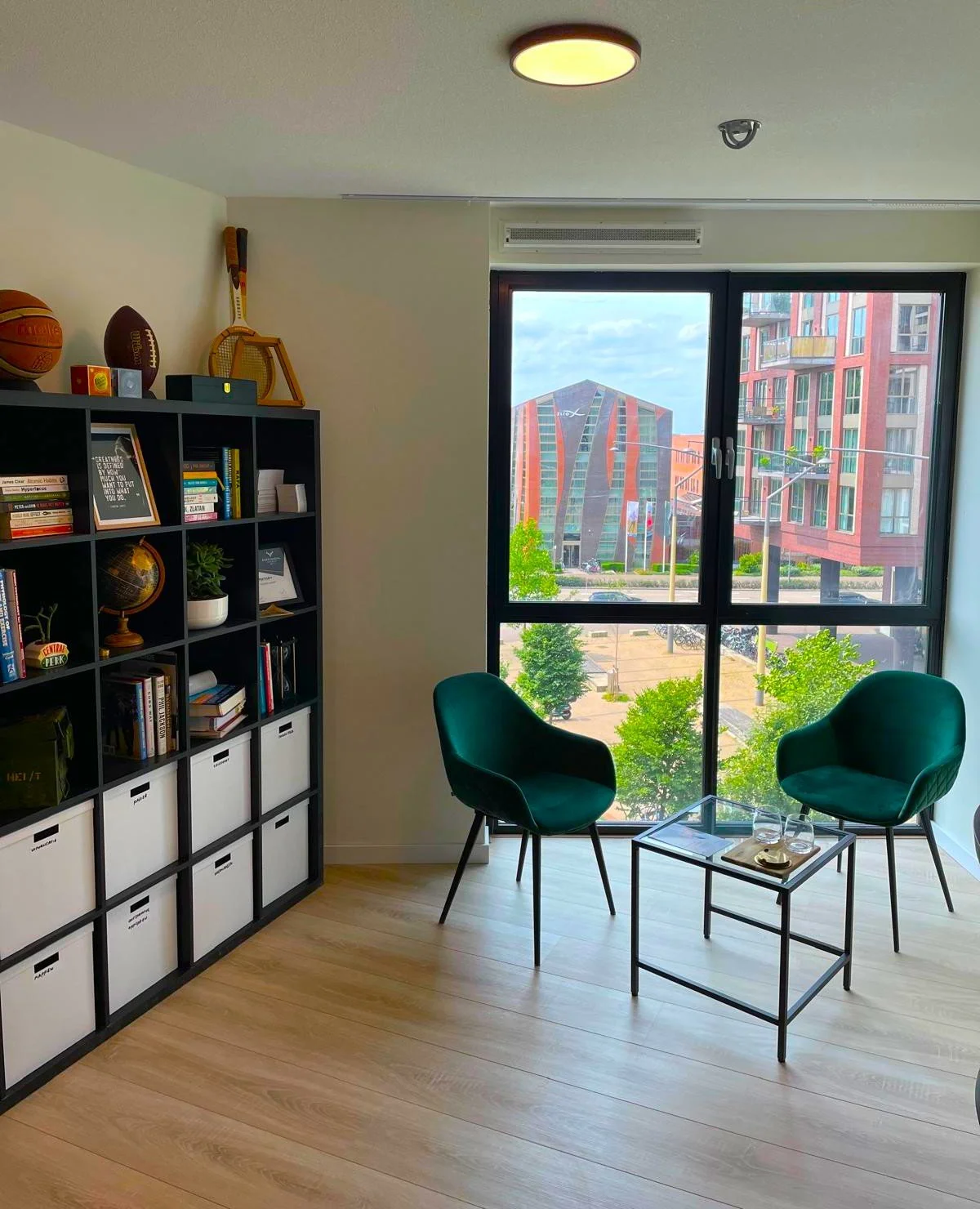 Interieur van een moderne woonkamer met twee groene stoelen, een glazen salontafel, een grote raam met uitzicht op kleurrijke gebouwen en groene bomen, en een zwarte boekenkast gevuld met boeken, decoraties, en opslagdozen.