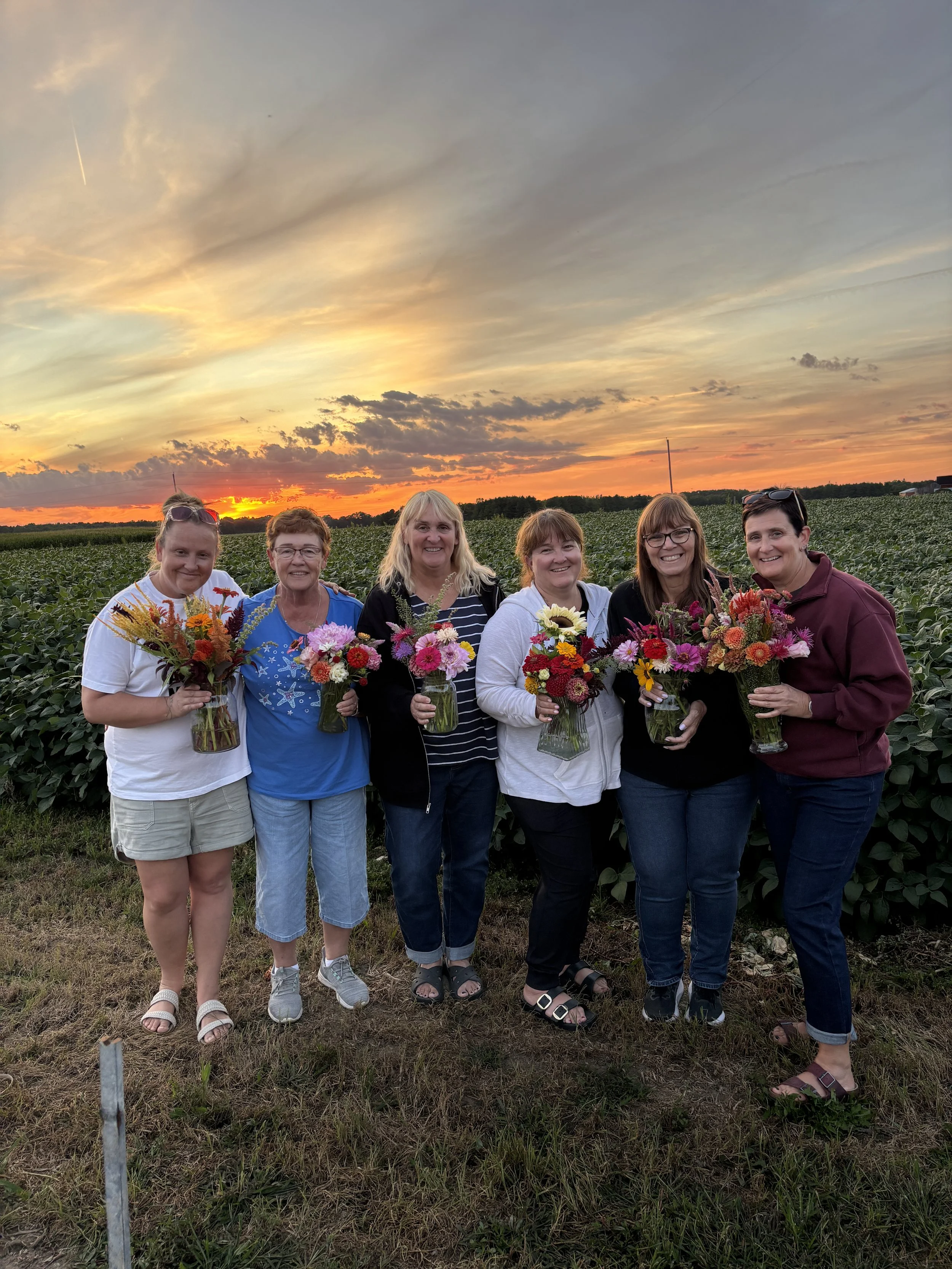 Six women standing in a field of crops during sunset, holding colorful flower bouquets and smiling.