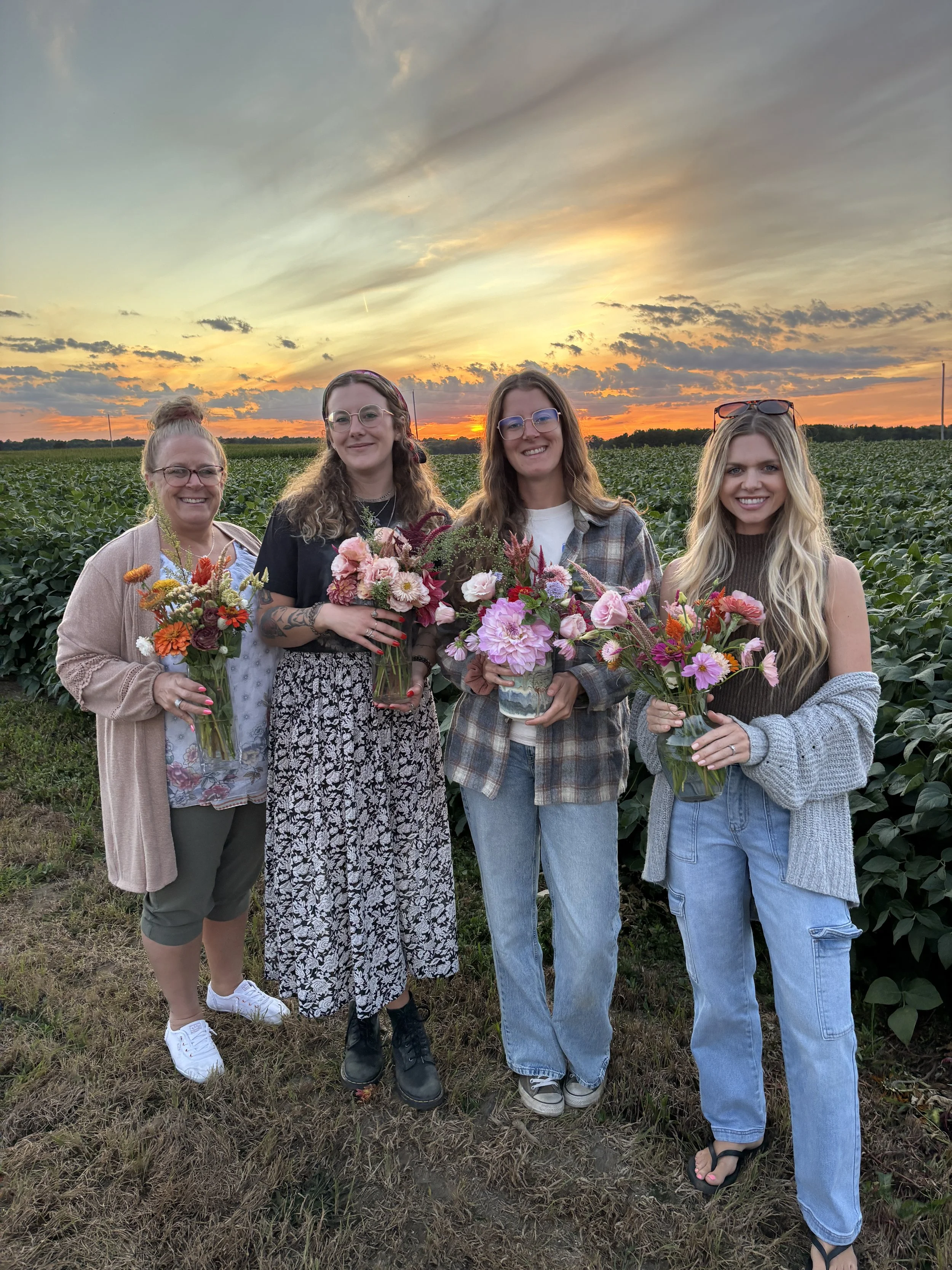 Four women standing in a field holding colorful flowers at sunset.