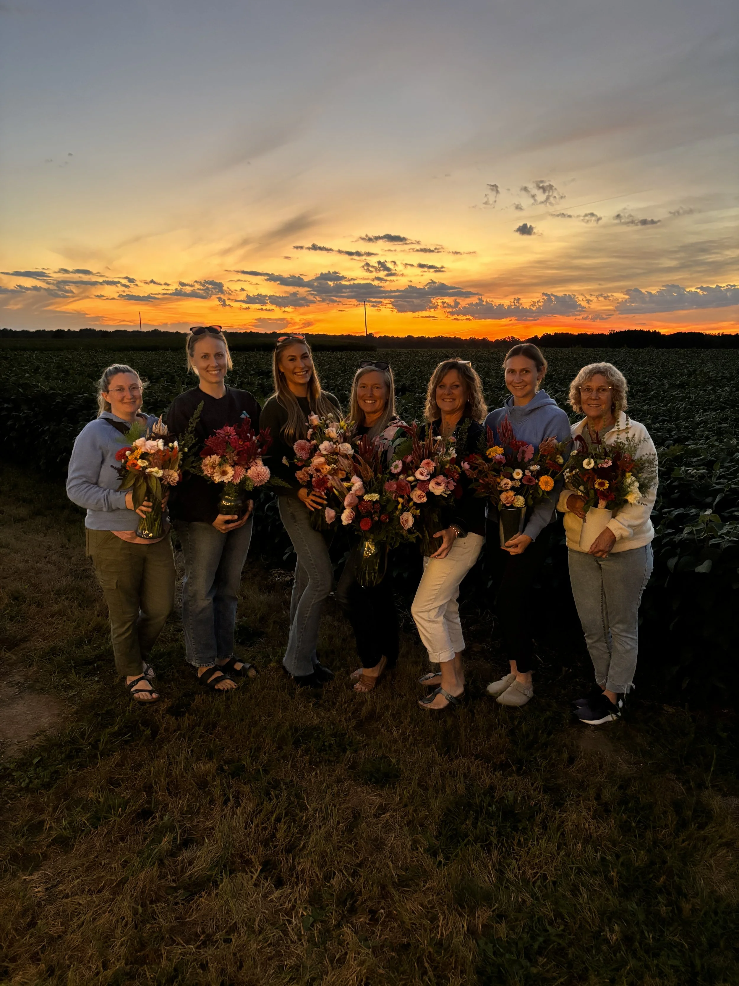 Seven women holding colorful flower bouquets standing in a field during sunset, with a sky filled with clouds and orange hues.