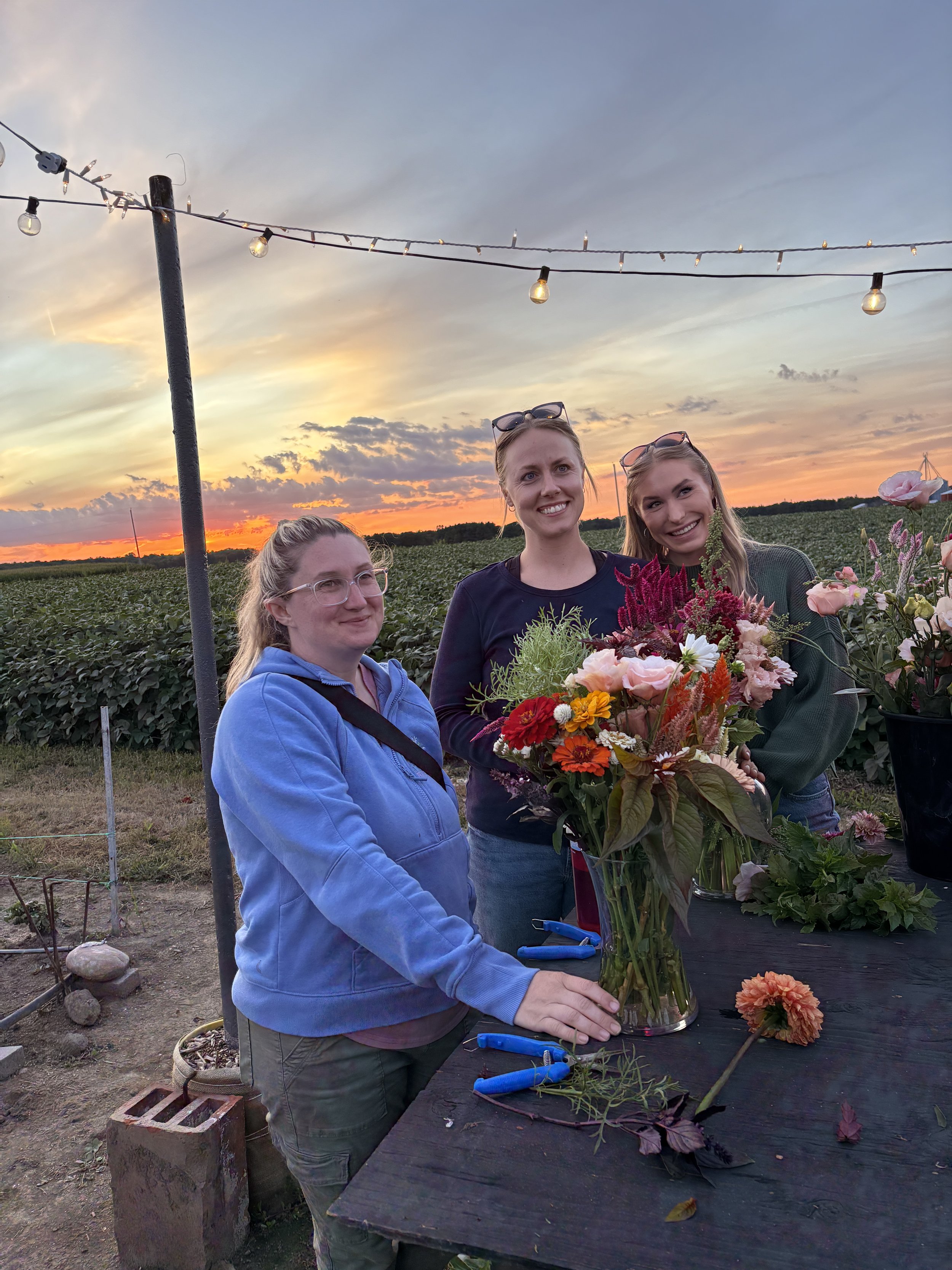 Three women standing outside at sunset, arranging flowers in a glass vase on a black table, with a field and a sunset sky in the background.