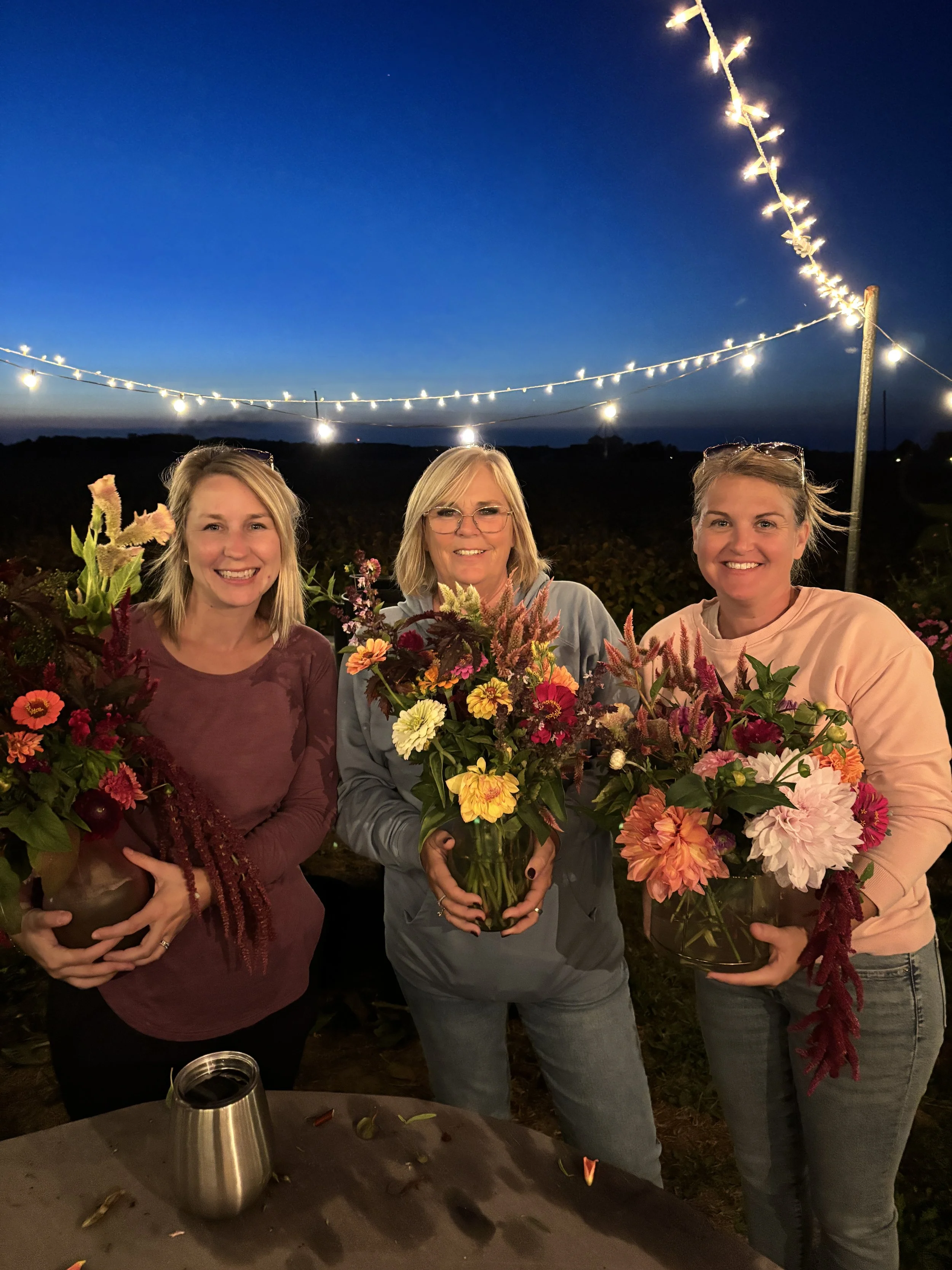 Three women smiling at night, holding colorful flower bouquets, with string lights hanging overhead and a dark sky in the background.