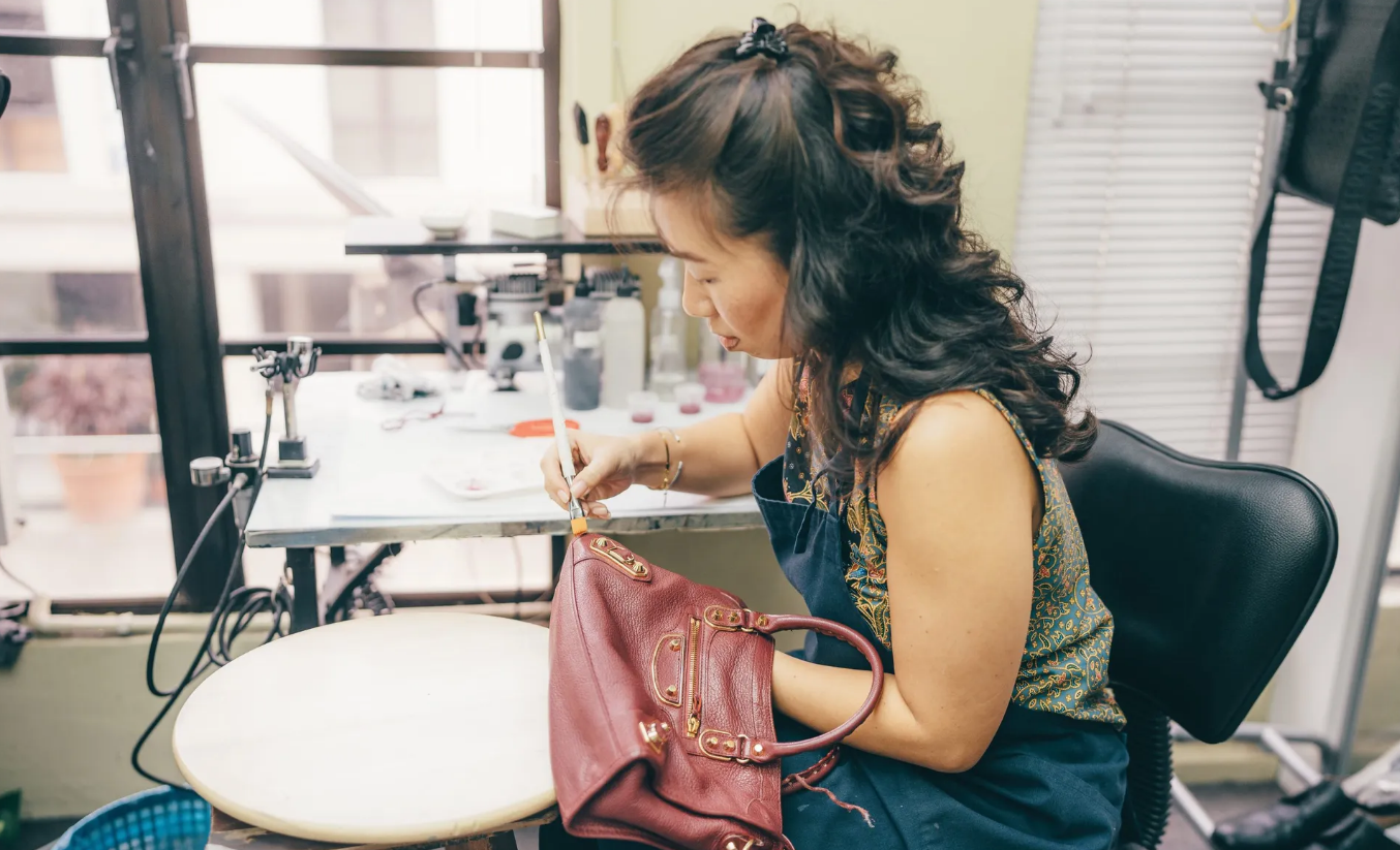 A woman working on a leather handbag in a workshop, sitting at a worktable with tools and materials, with a window and a shelf in the background.