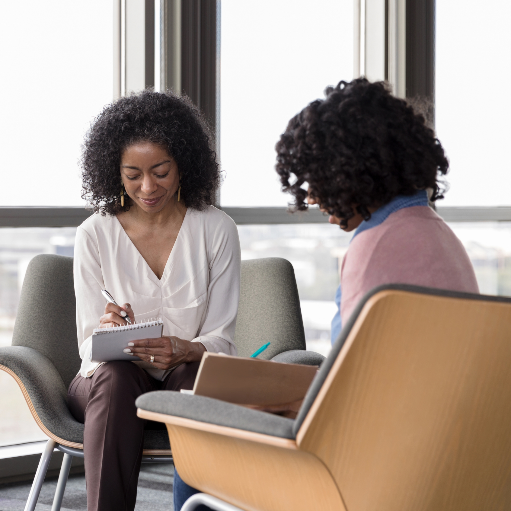 Four women with closed eyes embracing each other, wearing light pink attire, standing against a beige background.