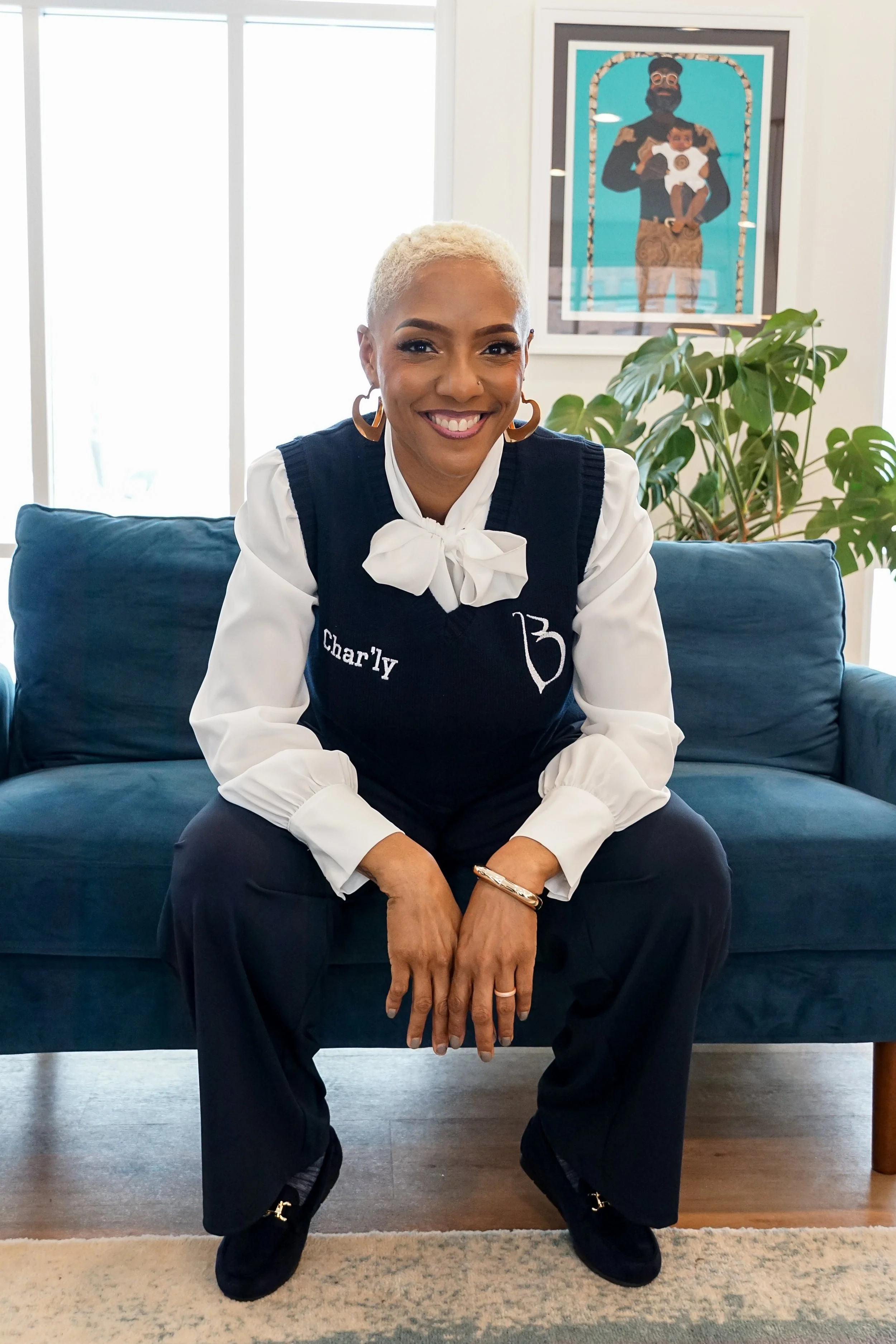 A smiling woman with short purple hair, wearing a black graphic T-shirt and a plaid blazer, standing with her arms crossed against a plain light background.
