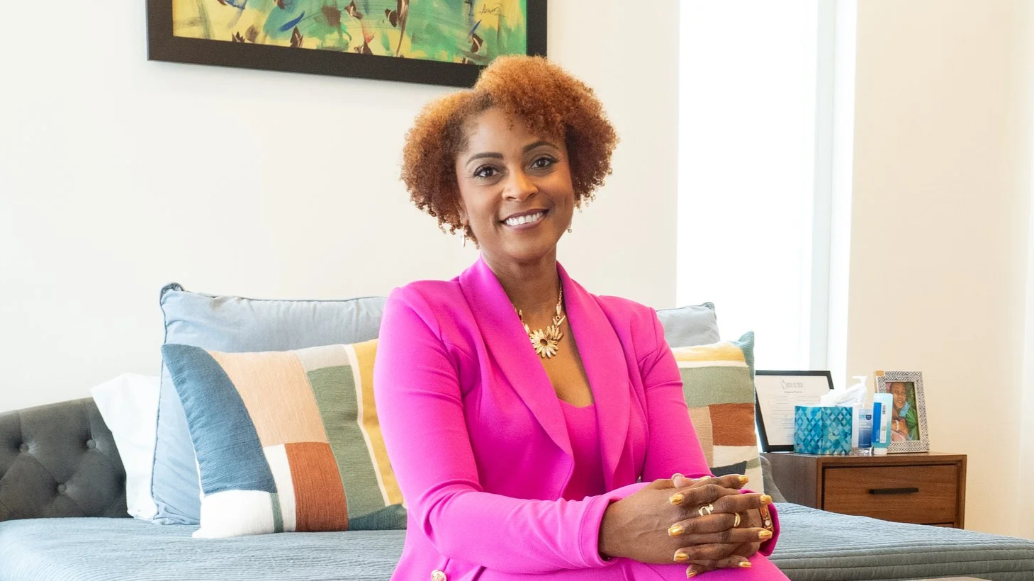 A woman with short curly hair wearing an orange blazer and white top, smiling at the camera, with hoop earrings and a light background.