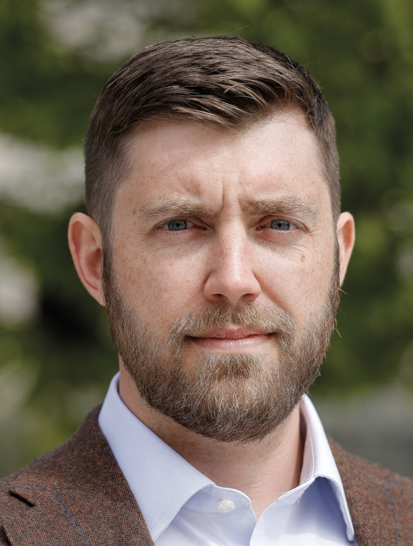 Close-up portrait of a middle-aged man with a beard and short brown hair, wearing a light blue shirt and a brown blazer, outdoors with blurred green trees in the background.