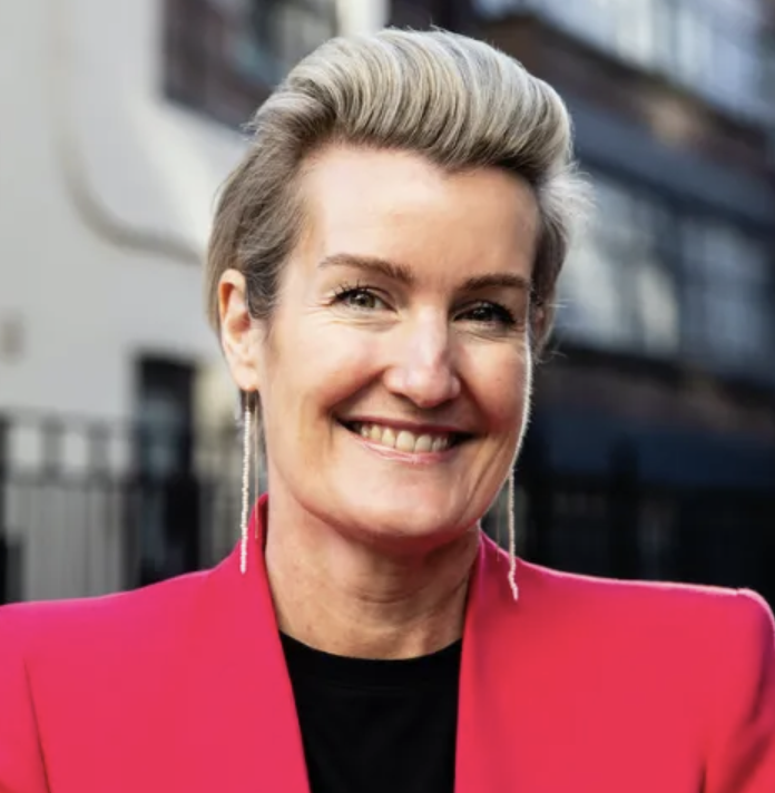 A smiling woman with short, styled blonde hair wearing a bright pink blazer and long earrings, standing outdoors in front of modern buildings.