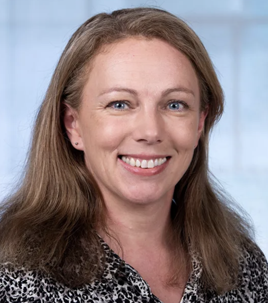 A woman with shoulder-length reddish-brown hair, blue eyes, and fair skin, smiling at the camera. She is wearing a patterned blouse and small earrings, with a soft, light-colored background.