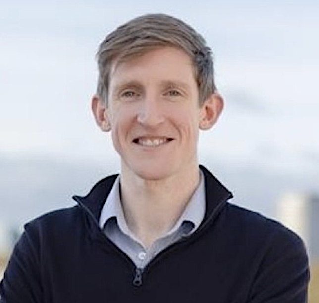 A smiling man with short light brown hair wearing a black zip-up jacket over a collared shirt, outdoors with a cloudy sky in the background.
