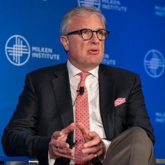 Man with gray hair and glasses, wearing a dark suit, white shirt, pink patterned tie, and pink pocket square, sitting in front of a blue background with the Milken Institute logo, speaking at an event.