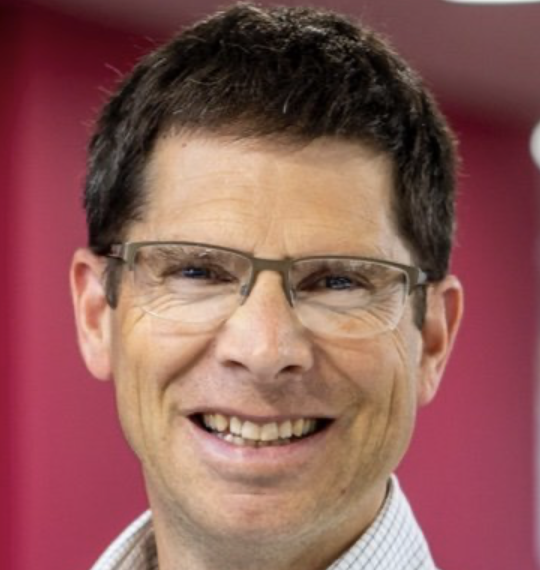 Close-up of a smiling man with glasses and short dark hair, wearing a light-colored shirt against a pink background.