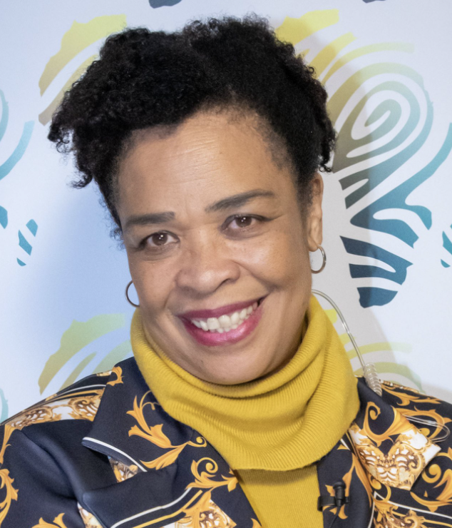 A woman with curly short hair, wearing a yellow turtleneck and a navy blazer with gold patterns, smiling in front of a colorful abstract background featuring stylized footprints.