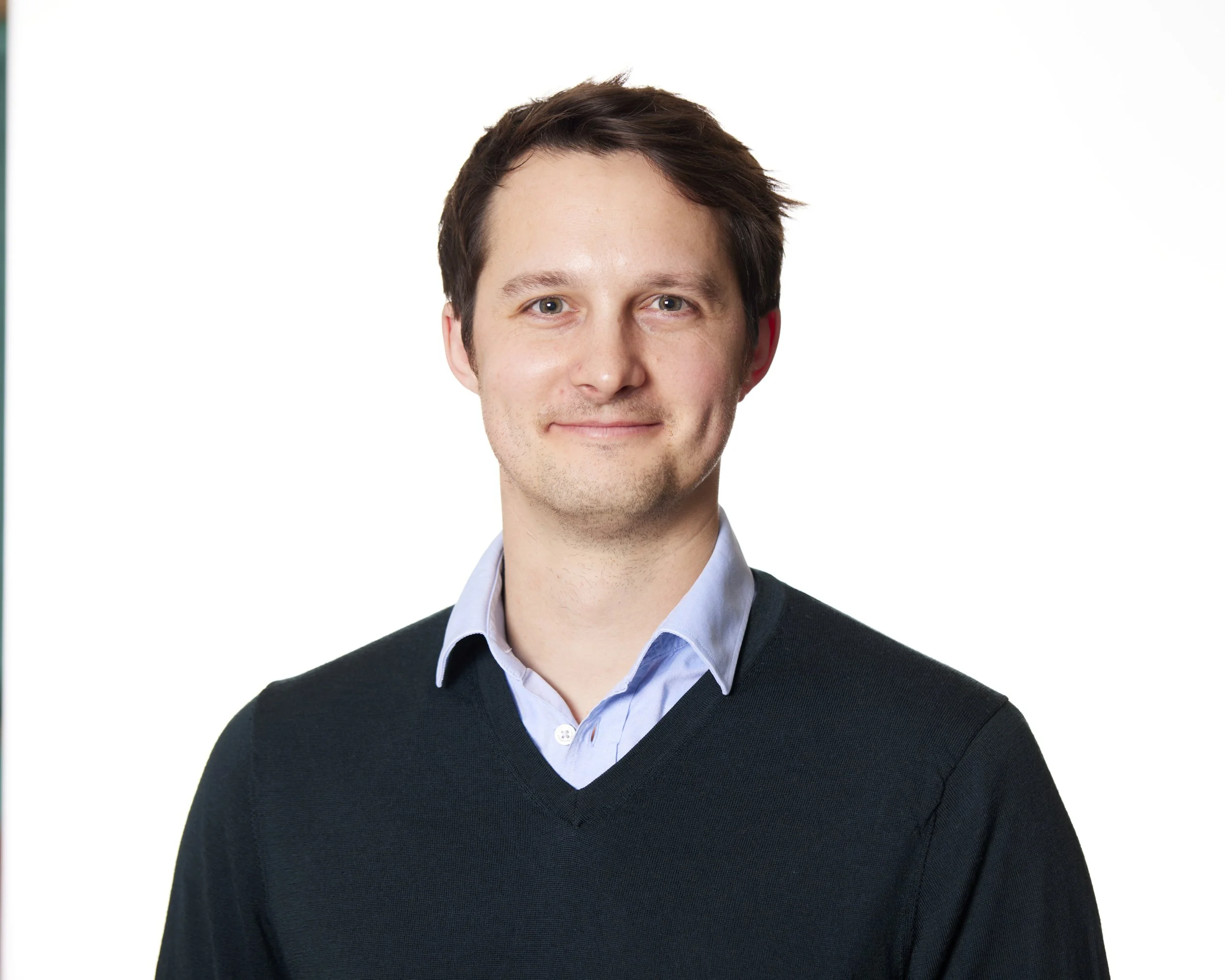 Portrait of a young man with brown hair, wearing a light blue collared shirt and a dark sweater, smiling against a white background.