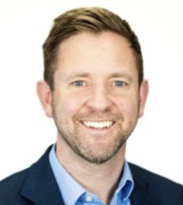 A smiling man with short brown hair and a beard, wearing a blue suit jacket and light blue shirt, against a white background.