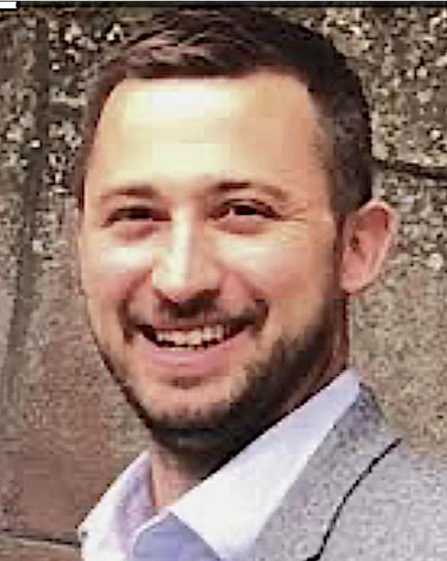 A smiling man with short dark hair and a beard, dressed in a blazer and shirt, standing outdoors in front of a tree.