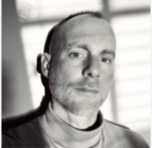 Black and white photo of a man with short hair and a slight beard, looking at the camera.