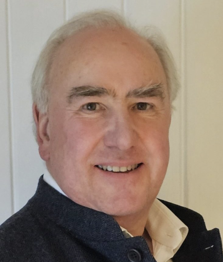A middle-aged man with gray hair smiling at the camera, wearing a dark sweater over a collared shirt, standing in front of a light-colored door.